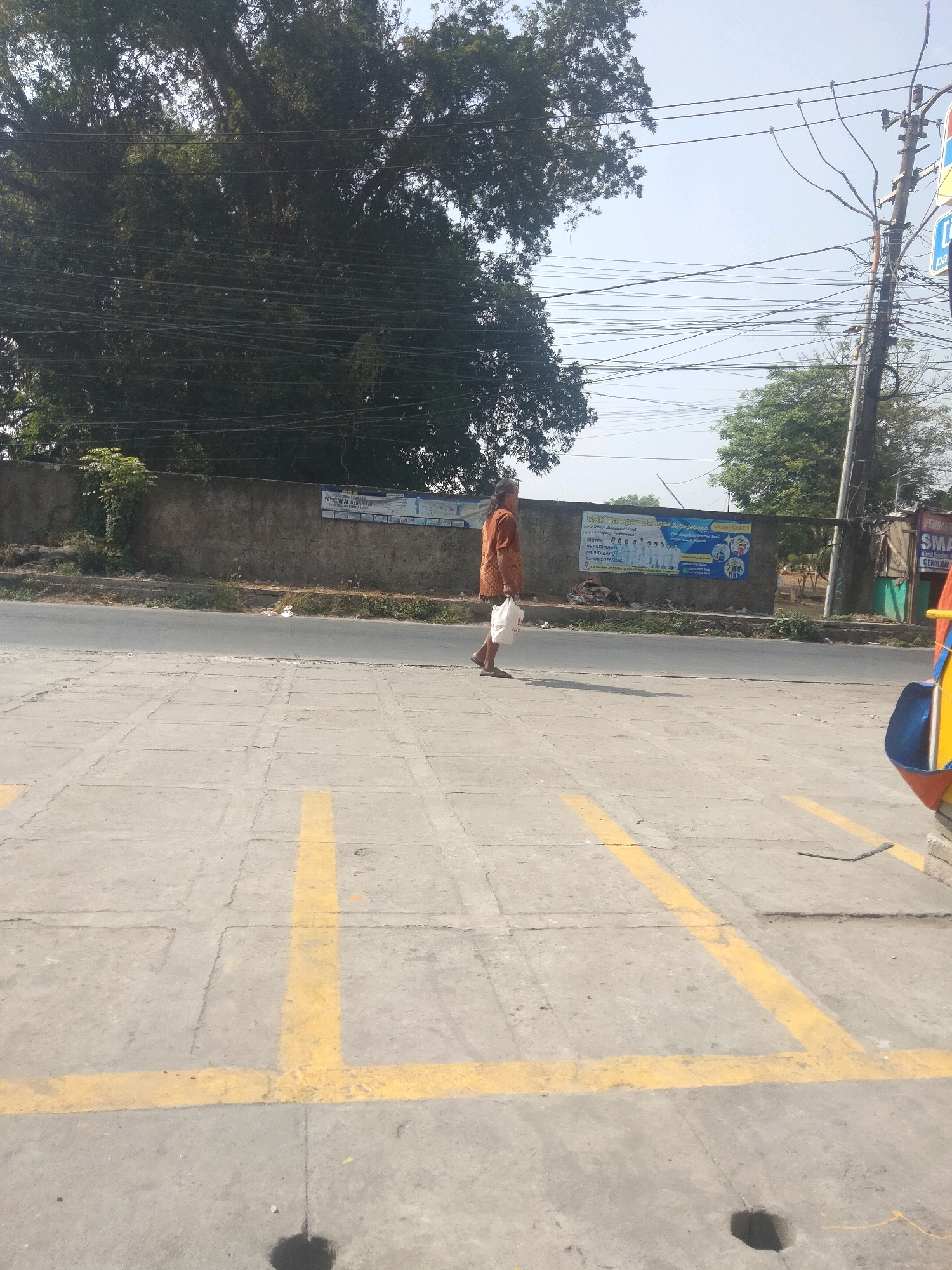 In Pisangan Jaya, Indonesia, a person walks along a paved walkway adjacent to a road. The individual, seen from the side, wears a light brown long-sleeved shirt and light-colored pants, carrying a white plastic bag in their left hand. They are walking away from the viewer, towards the left of the frame. The walkway is marked with yellow lines, creating a grid-like pattern. Behind the walkway, a low concrete wall runs parallel to the road. On the wall, a banner with text and images is displayed, featuring a school logo and photographs. Beyond the wall, a dense canopy of trees dominates the background, with electrical wires crisscrossing in front of them and a utility pole visible on the right. The sky above is a pale blue. The overall scene depicts a street view in a semi-urban or suburban area.