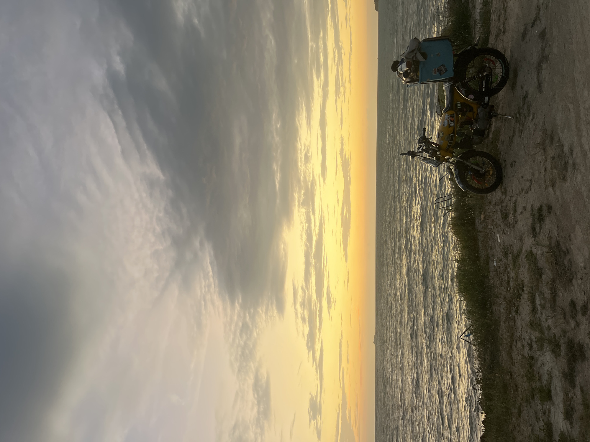 A yellow motorcycle with a light blue storage box filled with items is parked on a dirt path overlooking the ocean. The motorcycle has stickers and some visible damage. The ocean is calm with gentle waves, and the sky is filled with dramatic clouds, transitioning from a bright yellow and orange sunset near the horizon to darker gray clouds higher up. In the distance, a small island is visible on the horizon. The foreground shows sparse vegetation and dry earth. The scene appears to be a scenic overlook on the coast of Ko Pha-ngan District, Thailand, likely during sunset.