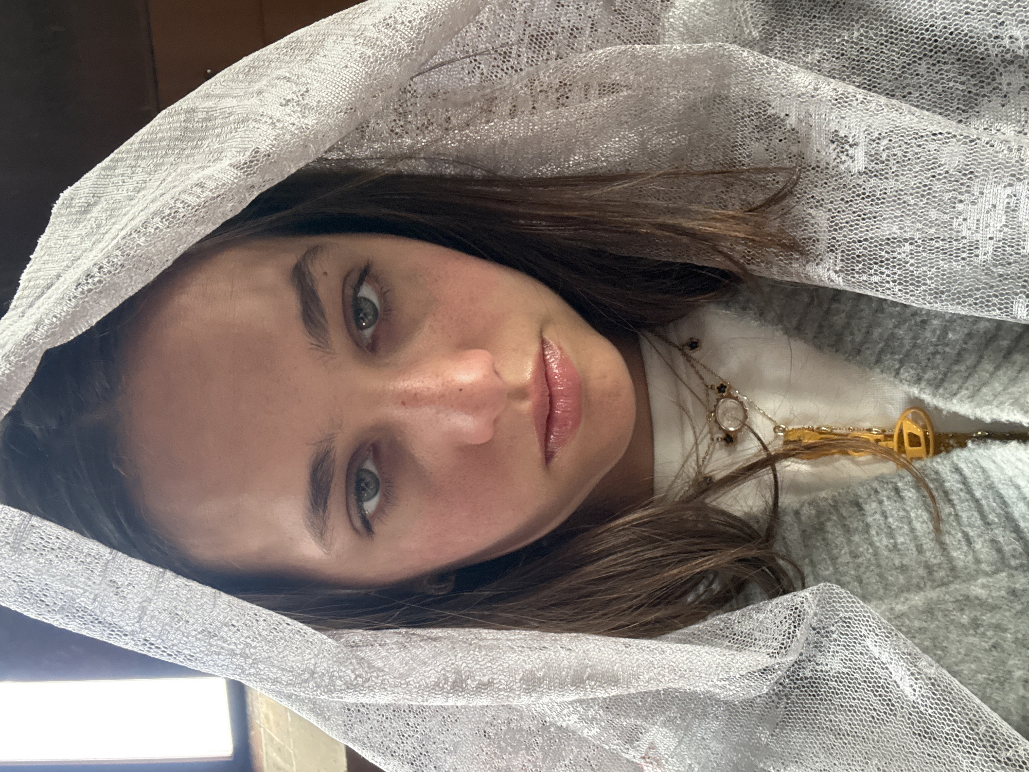 A woman is photographed indoors with a delicate, white, lace-like veil draped over her head and shoulders. Her face is tilted slightly. She has brown hair, fair skin with some freckles, and green eyes. She is wearing a light-colored shirt, a grey sweater, and a necklace with a gold key pendant. A window is visible in the lower left corner of the frame. It is impossible to determine the location within the city of Yerevan, Armenia, from the provided image.