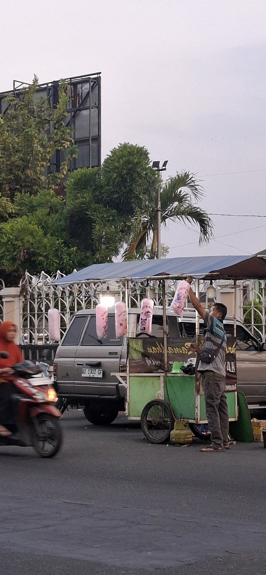 A person is standing next to a green food cart on a paved road. The food cart has a blue awning and is decorated with pink bags of cotton candy hanging from it. The person is holding one of the pink bags. Behind the food cart is a grey minivan with license plate AD 1302 GF. To the left of the minivan, a person on a red motorcycle is riding past. In the background, there are trees, a white fence, and a large billboard structure. The scene appears to be in a street in Desa Donohudan, Indonesia, given the Indonesian license plate and the overall environment. The sky is overcast.