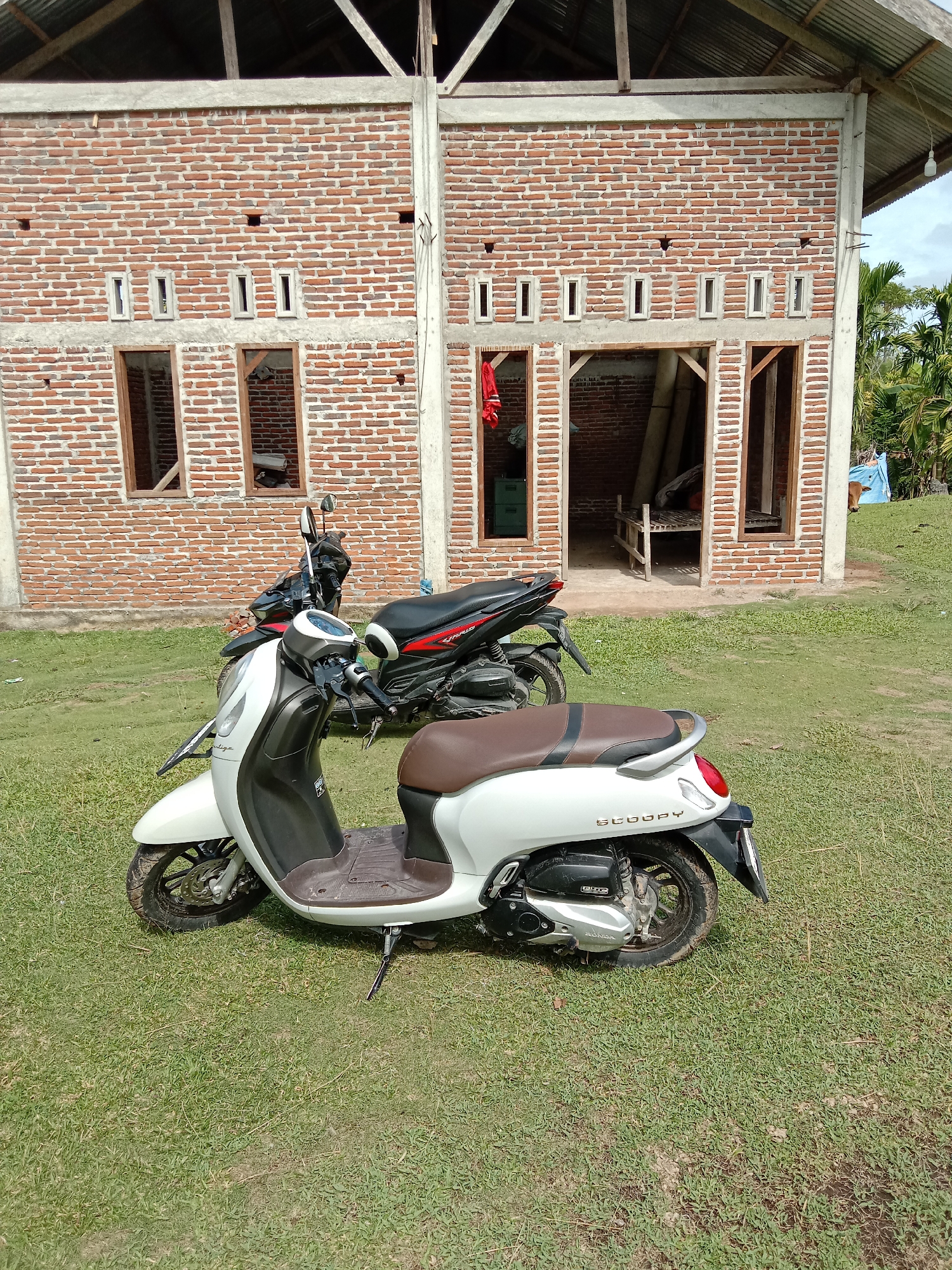 The image shows a Scoopy motorcycle parked on a grassy field in front of a brick building with window and door openings. Another motorcycle is partially visible behind the Scoopy. The environment appears to be a rural area, with trees visible in the background. The location is Blangkarieng, Indonesia.
