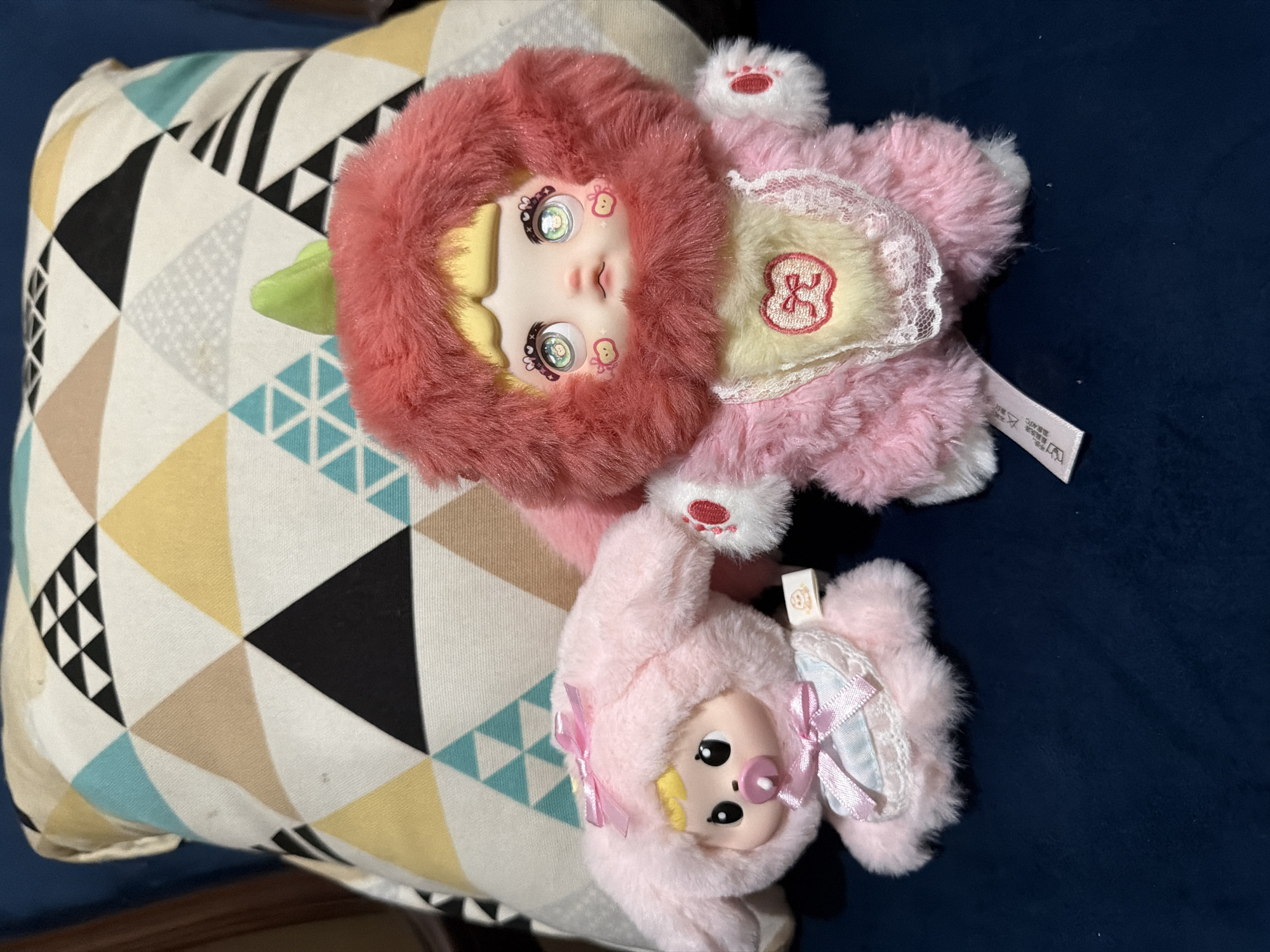 The image shows two pink plush dolls, one larger and one smaller, lying on a patterned cushion which in turn is resting on a blue surface. The larger doll has a red fuzzy hood and a pale face with unique eye details and markings on the cheeks. It has a yellow patch on its chest decorated with a red bow symbol and surrounded by lace. The smaller doll has a pink fuzzy hood with pink bows and a pacifier. The background is a solid blue color. There is no information in the image to confirm the location in East Jakarta, Indonesia.