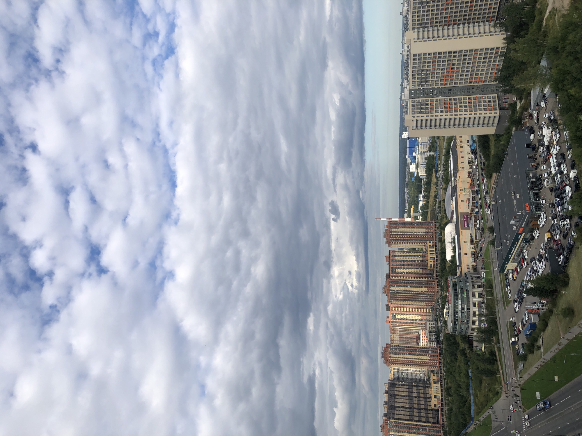 A high-angle shot captures a sprawling urban landscape under a cloudy sky. The scene is dominated by rows of tall, modern apartment buildings with colorful facades, predominantly in shades of orange, yellow, and beige. A large, circular building with glass windows is visible in the mid-ground, surrounded by lush green trees. Adjacent to this is a large commercial building with a prominent sign reading "LADA" above a car dealership lot filled with vehicles. Several roads are present, with cars and a blue bus navigating through them. In the distance, more industrial buildings and a tall red and white striped smokestack can be seen. The sky is a mosaic of dark gray and bright white clouds, suggesting an overcast or partially sunny day. The overall impression is of a modern residential and commercial district.