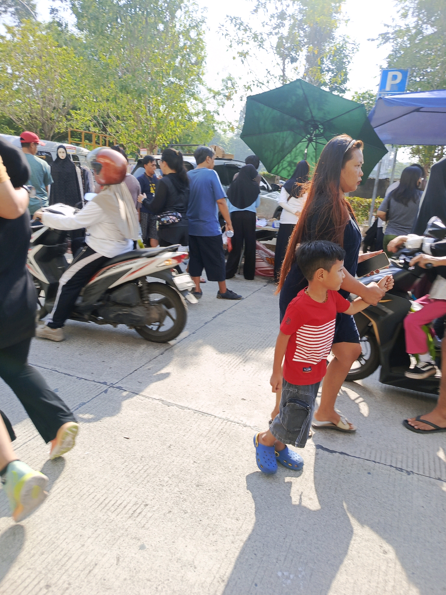 The image depicts a street scene in Tangerang, Indonesia, likely a marketplace or roadside area based on the presence of stalls and a diverse group of pedestrians and vehicles. The sun is bright, casting strong shadows, indicating daytime.
In the foreground, a young boy in a red t-shirt with white stripes and denim shorts walks to the right, wearing blue Crocs. To his right, a woman with long, dark hair, wearing a dark blue dress, holds his arm. She is walking to the right, with a black smartphone in her hand. Behind them, a black motorcycle with a passenger in pink pants is visible.
Further back, a person on a white and black scooter is moving to the left. This individual is wearing a white shirt and a light-colored head covering, with a red helmet. To the left of the scooter, a person in a black outfit is walking away from the camera. In the background, several other people are present, some under awnings or umbrellas, suggesting a commercial or gathering area.
Notable objects include a large green umbrella, a blue awning with a parking sign (P), and various vehicles, including scooters and possibly cars partially obscured. The ground is paved with concrete, showing lines and cracks. Trees with dense green foliage are visible in the background, typical of a tropical environment. The overall atmosphere appears to be one of moderate activity, possibly a busy public space in Tangerang.