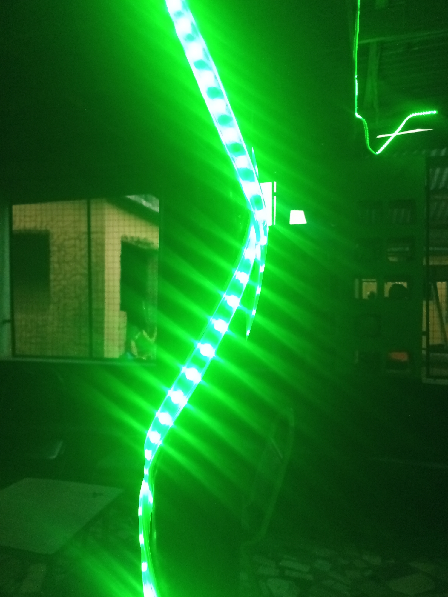 An overhead shot shows a coiled green LED light strip with white LEDs in a dimly lit indoor space. The light strip is draped across the frame, creating a diagonal line from the bottom left to the top right. The light from the strip casts bright green beams and reflections on the dark surroundings. In the background, to the left, is a window with a grid pattern revealing a blurred interior or exterior with beige walls. To the right, more of the dimly lit environment is visible, with dark furniture or structures and some lighter colored objects that are indistinct due to the low light and green glow. The overall scene appears to be a room or an open space with furniture and architectural elements.