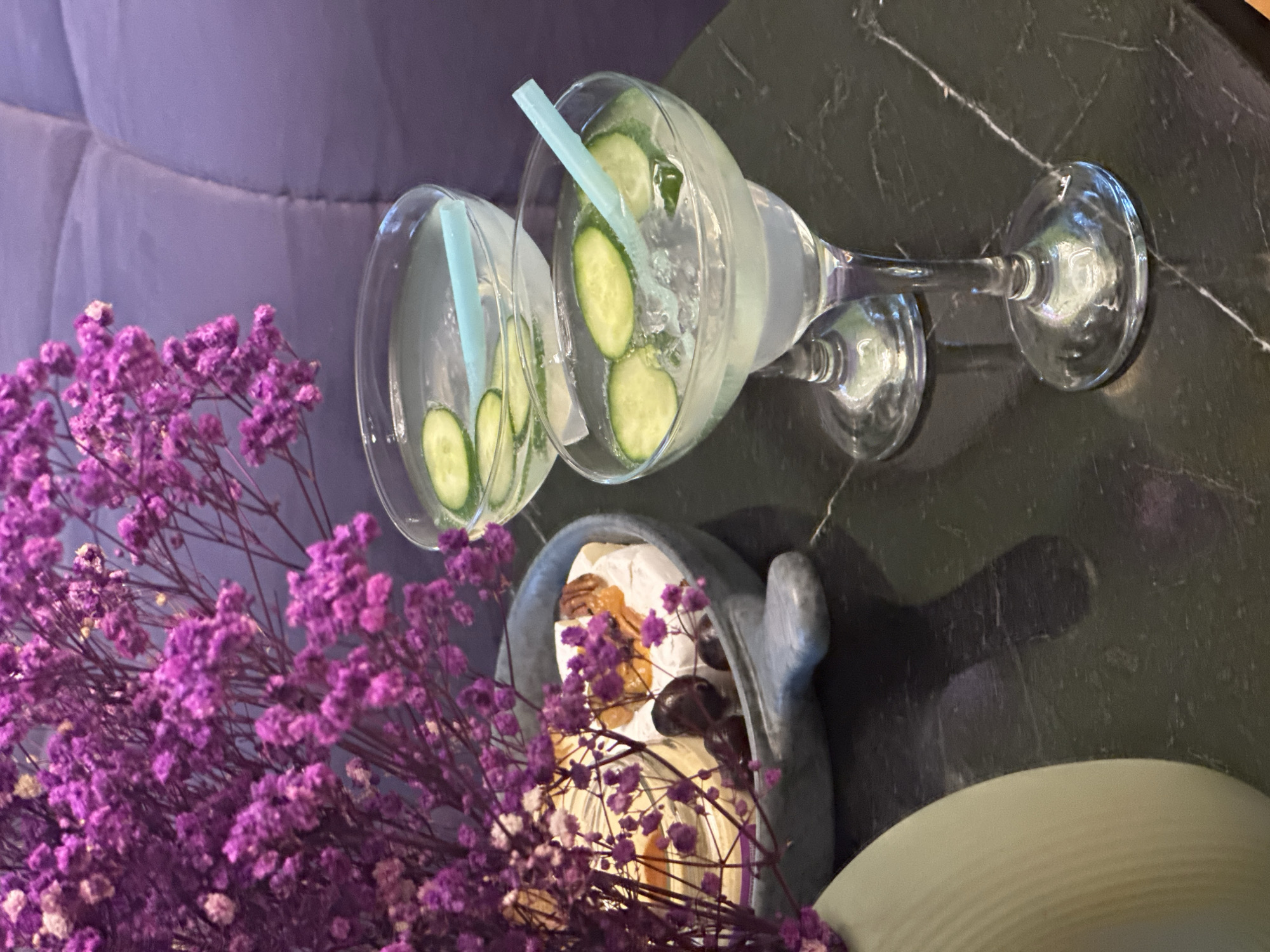 In the foreground, a vibrant bouquet of small, clustered purple flowers with thin, dry branches adds a splash of color. Behind the flowers, two glasses of clear liquid with ice and cucumber slices are prominently displayed. Both glasses contain light blue straws. One glass is a coupe-style glass, while the other is a wider, shallow glass, possibly a martini glass. The drinks appear to be refreshing, likely cocktails or infused water.
Further back, a grey, bowl-like object is visible, containing what appears to be a pastry or dessert with nuts and a glossy topping. This object is positioned on a dark, speckled surface, likely a marble tabletop. In the background, a plush, padded surface in a soft purple hue suggests a comfortable seating area, perhaps a couch or chair.
The lighting suggests an indoor setting, possibly in the late afternoon or evening, as the light is soft and diffused. The overall mood is tranquil and inviting, with a focus on refreshments and decorative elements. There is no visible text in the image. The context of Novosibirsk, Russia, does not provide specific visual cues within the image itself, but it sets a geographical backdrop.