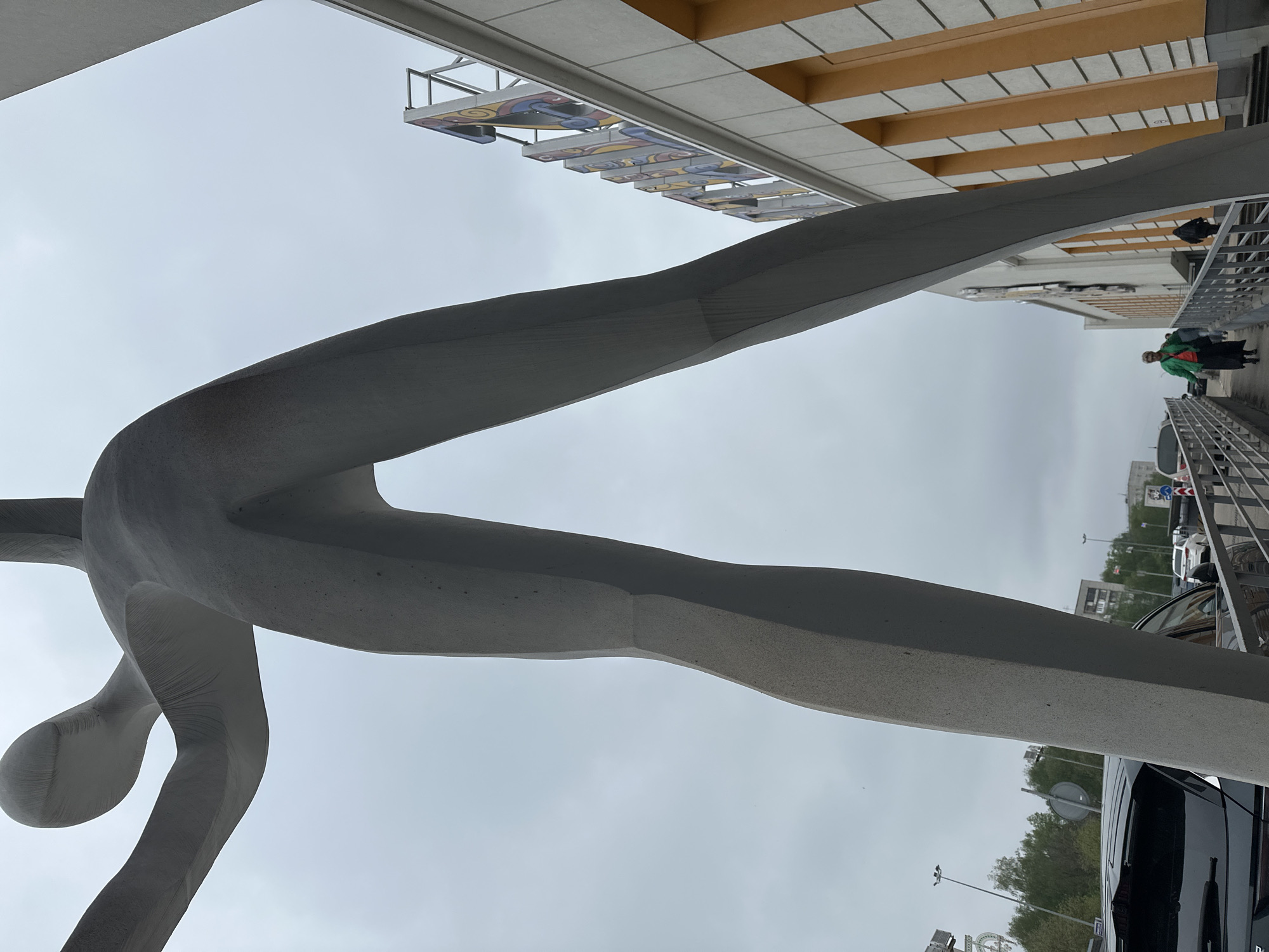 The image shows a concrete sculpture of a stylized human figure in an outstretched pose, possibly running or jumping. It is located in Nizhny Novgorod, Russia. The sculpture stands in front of a building with a facade of white and yellow panels, with a sign overhead featuring colorful, abstract designs. To the right of the sculpture is a pedestrian bridge with metal railings where two people are walking: one is wearing a green jacket, and the other is further away and less distinct. Below the bridge, a parking lot contains parked cars, including a black SUV. The sky is overcast.