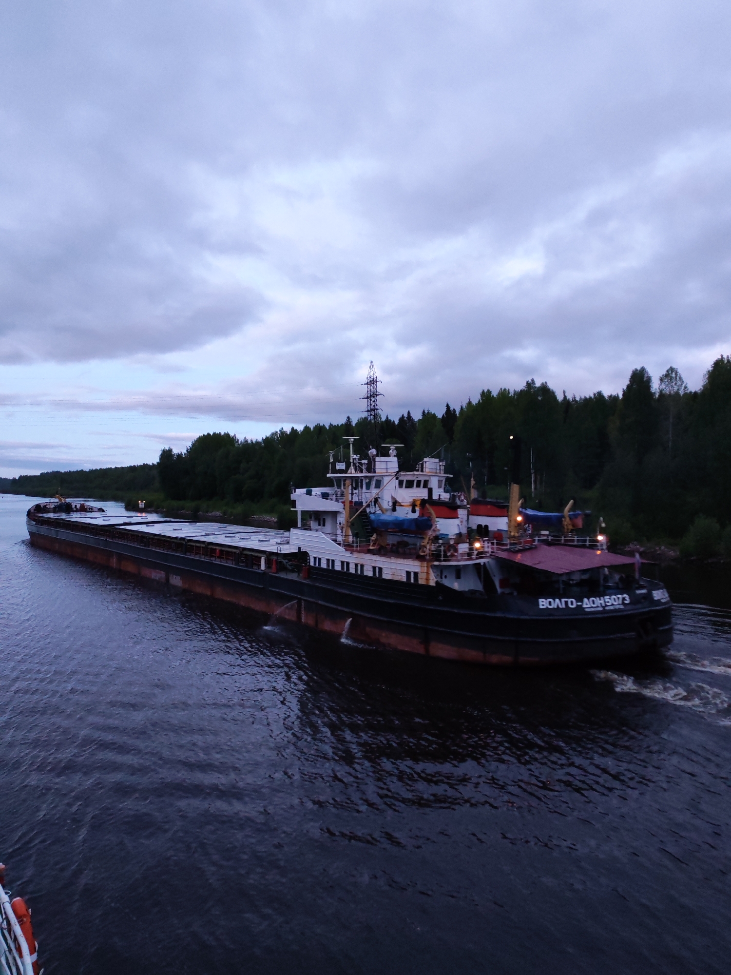 A large barge, identified as "ВОЛГО-ДОН 5073", is moving on a body of water. The barge is dark in color with a white superstructure. It appears to be passing through a canal or narrow river, with trees lining both banks. An electrical transmission tower is visible in the background. The sky is overcast. There is no definitive proof the image was taken in Александровское, Russia.