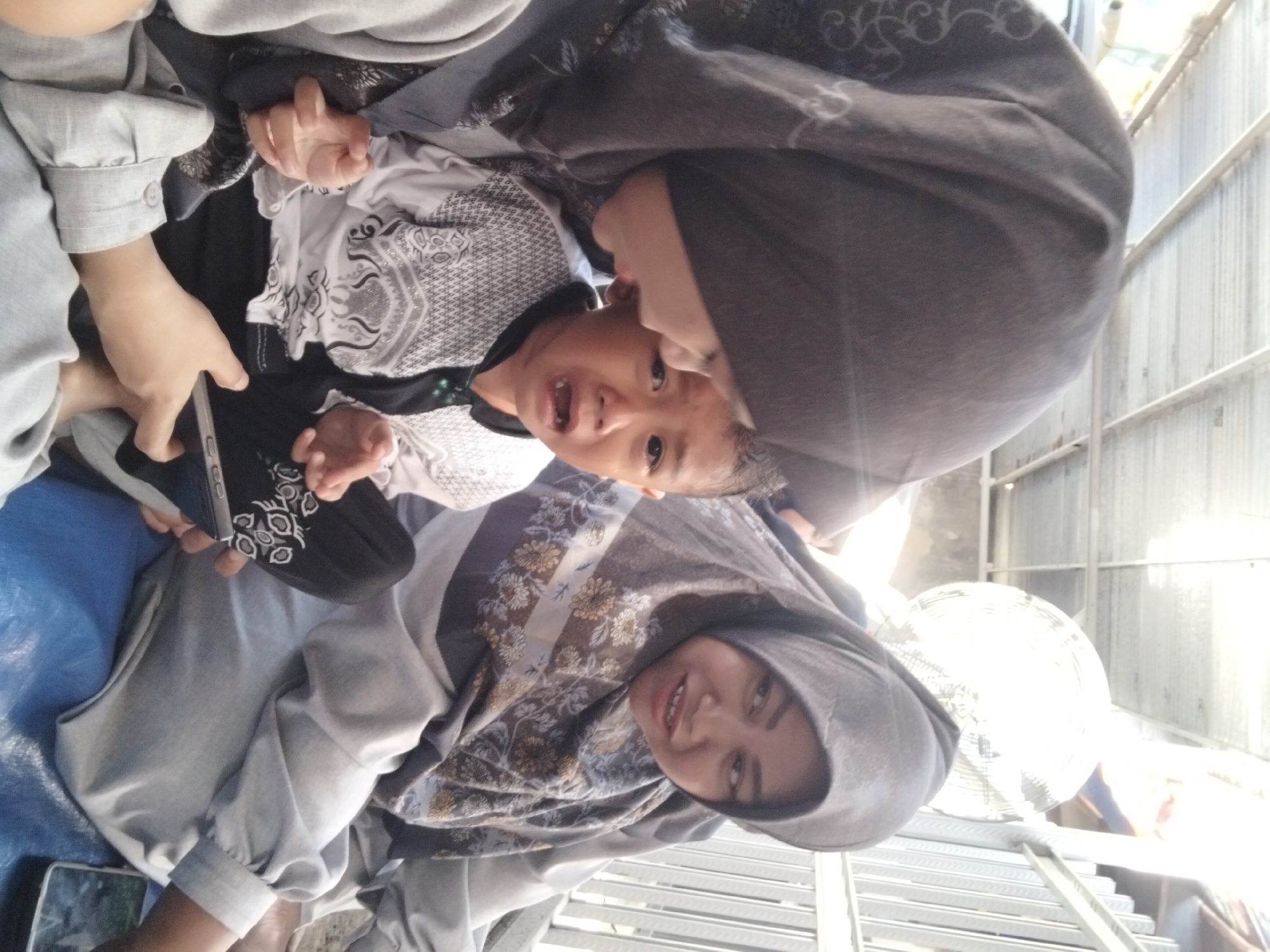 In the city of Kepuh Kiriman, Indonesia, two women and a crying child are captured in a close-up, outdoor photograph. The scene appears to be under a corrugated metal roof, suggesting an open-air shelter or a covered walkway. The woman on the left, wearing a dark hijab with a subtle pattern, leans in towards the child. Her arm is around the child, and she holds a smartphone in her hand, possibly showing it to the child. The child, wearing a white shirt with a black collar and intricate grey and white patterned sleeves, is crying intensely, with mouth open and tears visible. The woman on the right, wearing a hijab with a floral pattern in shades of gray and brown, looks away from the camera with a slight smile. Her body is angled towards the child and the other woman. The child's hands are visible, one with fingers spread open. The environment includes a metal railing in the background and a white electric fan. The lighting suggests it is daytime, with bright sunlight.
