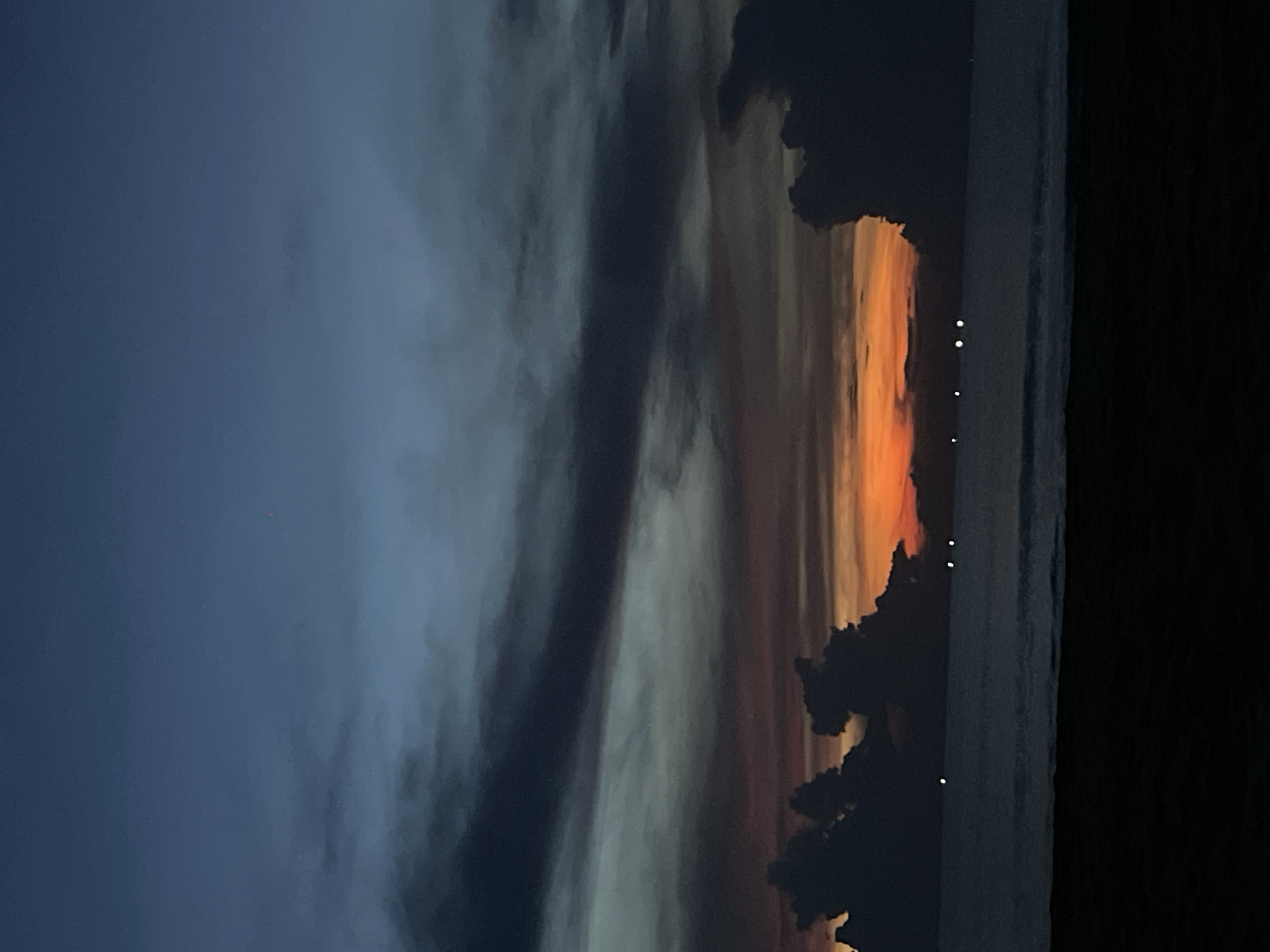 The image captures a dusky sky over a body of water, likely the ocean, with a beach in the foreground. The sky transitions from a deep blue at the top to shades of gray and orange near the horizon, indicating either dawn or dusk. Dark, heavy clouds are present, some silhouetted against the brighter sky. The water is calm with small waves visible, and there are a few distant lights, possibly from boats or a coastal city. The beach in the foreground is dark and out of focus, suggesting it is made of sand. The overall scene evokes a sense of tranquility and the transition between day and night.
While the image itself does not contain any specific visual cues to definitively place it in Da Nang, Vietnam, the prompt specifies this location. Da Nang is a coastal city in central Vietnam known for its long sandy beaches and vibrant waterfront. If this image were taken in Da Nang, it would likely be depicting one of its beaches, such as My Khe Beach or Non Nuoc Beach, during a twilight period. The presence of distant lights could be indicative of the city's urban skyline visible from the coast.