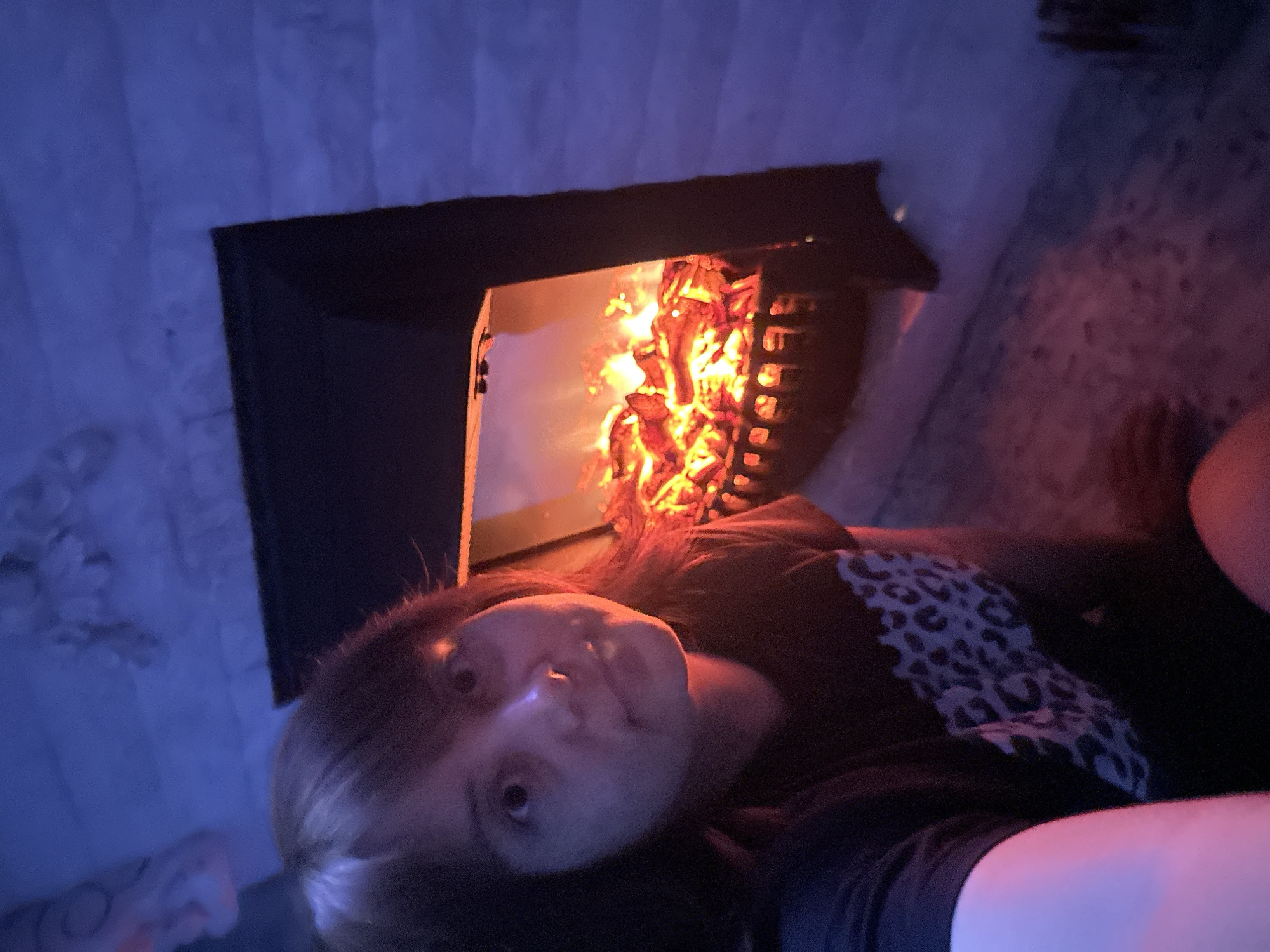 A person is taking a selfie in front of a fireplace. The person's face is in the foreground, illuminated by the fire's glow. The fireplace is black and filled with burning logs. The background is a stone wall bathed in blue light. The person is wearing a dark t-shirt with a leopard print design. The scene appears to be indoors, possibly in a home.