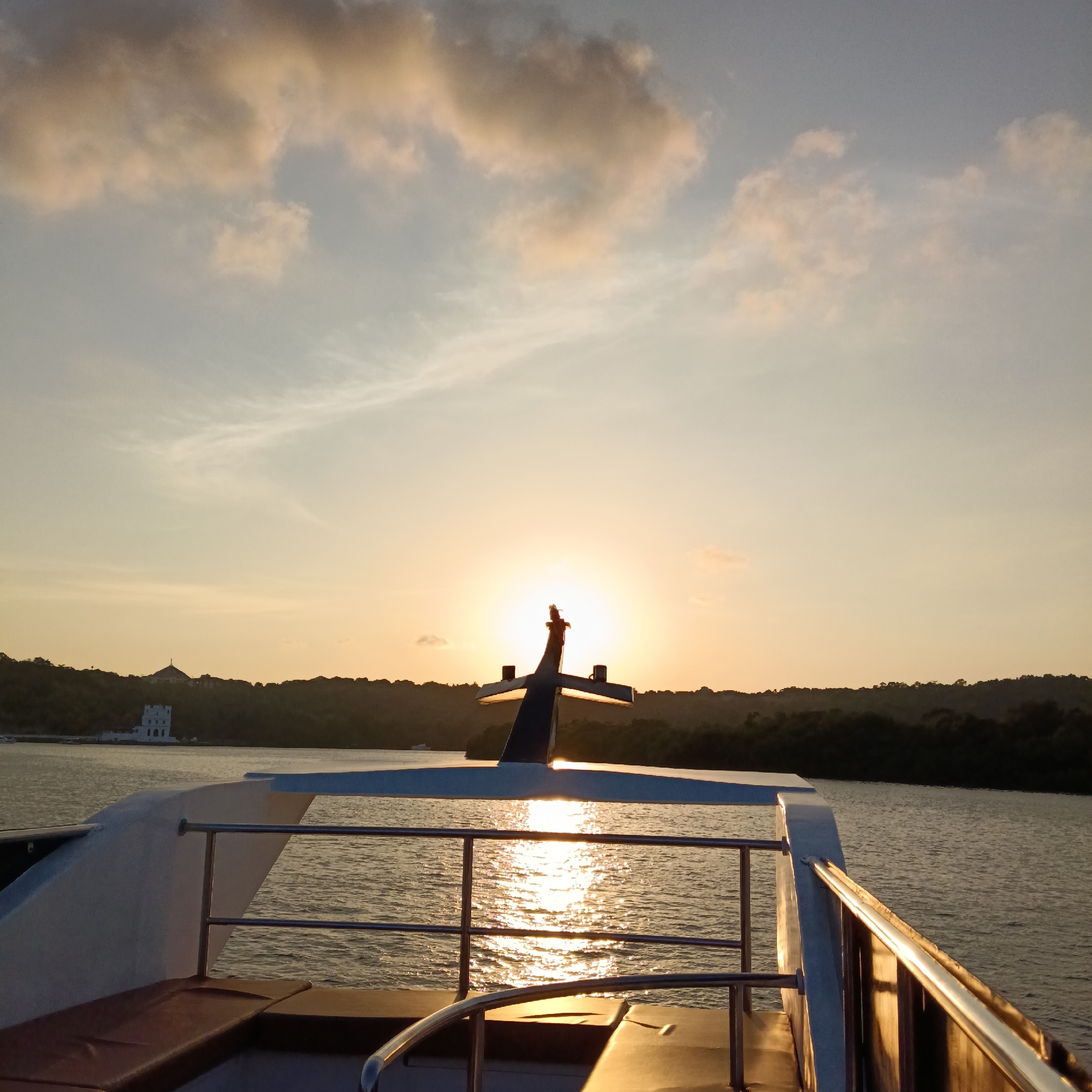 The image shows a view from the stern of a boat at sunset. The sun is centered and shining between the boat's mast or antenna and reflecting on the water. The railing is visible. The shoreline is in the background. A white building can be seen among the trees on the shoreline. It is likely the location is Britona, India.
