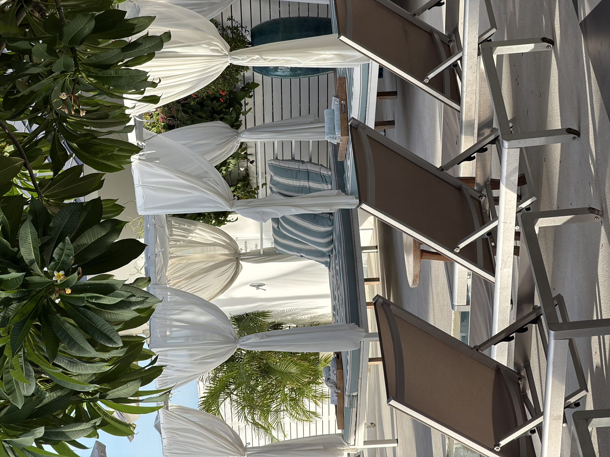 The image depicts a poolside lounging area in Baan Bo Phut, Thailand, featuring several empty lounge chairs and cabanas. In the foreground, three metallic lounge chairs with brown fabric are angled towards the viewer. Behind these chairs, a cabana with white draped fabric offers shade over a daybed. This daybed is adorned with striped cushions in blue and white, and a wooden tray holding neatly folded blue towels rests on its surface. Another daybed, also partially visible, is situated in the background. Lush green foliage, including palm fronds and flowering branches, hangs over the cabanas, casting dappled shadows on the ground. A tall, teal ceramic vase stands near the cabana on the right, and a white slatted wall forms the backdrop. The ground appears to be a textured concrete surface. The overall scene conveys a sense of serene, resort-style relaxation.