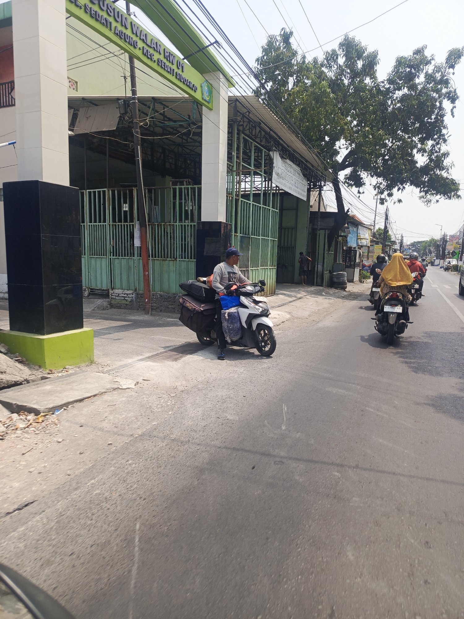 The image depicts a street scene in Sedati Agung, Indonesia. A man wearing a blue cap and a grey shirt is riding a white scooter laden with luggage. He is traveling on a paved road. To his left is a building with a green and white facade, displaying text that reads "DUSUN WALAN RW 01 SEDATI AGUNG KEC. SEDATI SIDOARJO". The building has a green metal fence in front of it, and a large tree with dense foliage partially obscures the upper portion of the scene. Other motorbikes are visible further down the road, some with passengers. The sky is overcast. The overall impression is of a busy street with pedestrian and vehicular traffic.