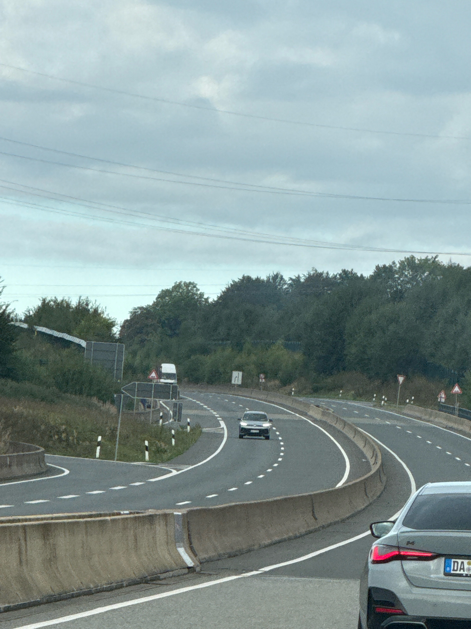 The image captures a highway in Germany under a cloudy sky. In the foreground, the rear of a light grey BMW sedan is visible, indicating the viewpoint is from another vehicle on the road. The highway curves to the right, with concrete barriers separating lanes. Several other vehicles are present on the highway, including a silver sedan further ahead and a white van on an exit ramp. The surrounding environment is characterized by dense green trees on either side of the road and overhead power lines stretching across the sky. Road signs indicating traffic direction and potential hazards are visible along the roadside. The license plate of the BMW is partially visible and reads "DA". This scene is consistent with a highway location in or near Bad Lauterberg im Harz, Germany, known for its scenic routes and wooded landscapes.