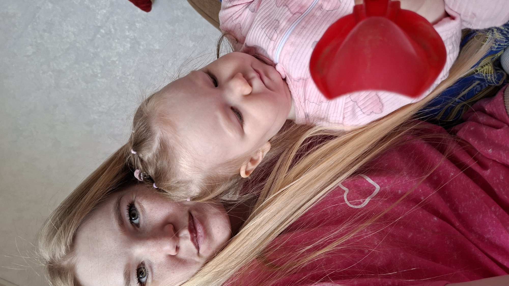 A woman and a baby are lying down. The woman has long blonde hair and is wearing a pink shirt with a white heart. The baby is wearing a pink onesie and has a red plastic toy nearby. The location is likely indoors. There is no evidence of Большой Букор or any urban environment.