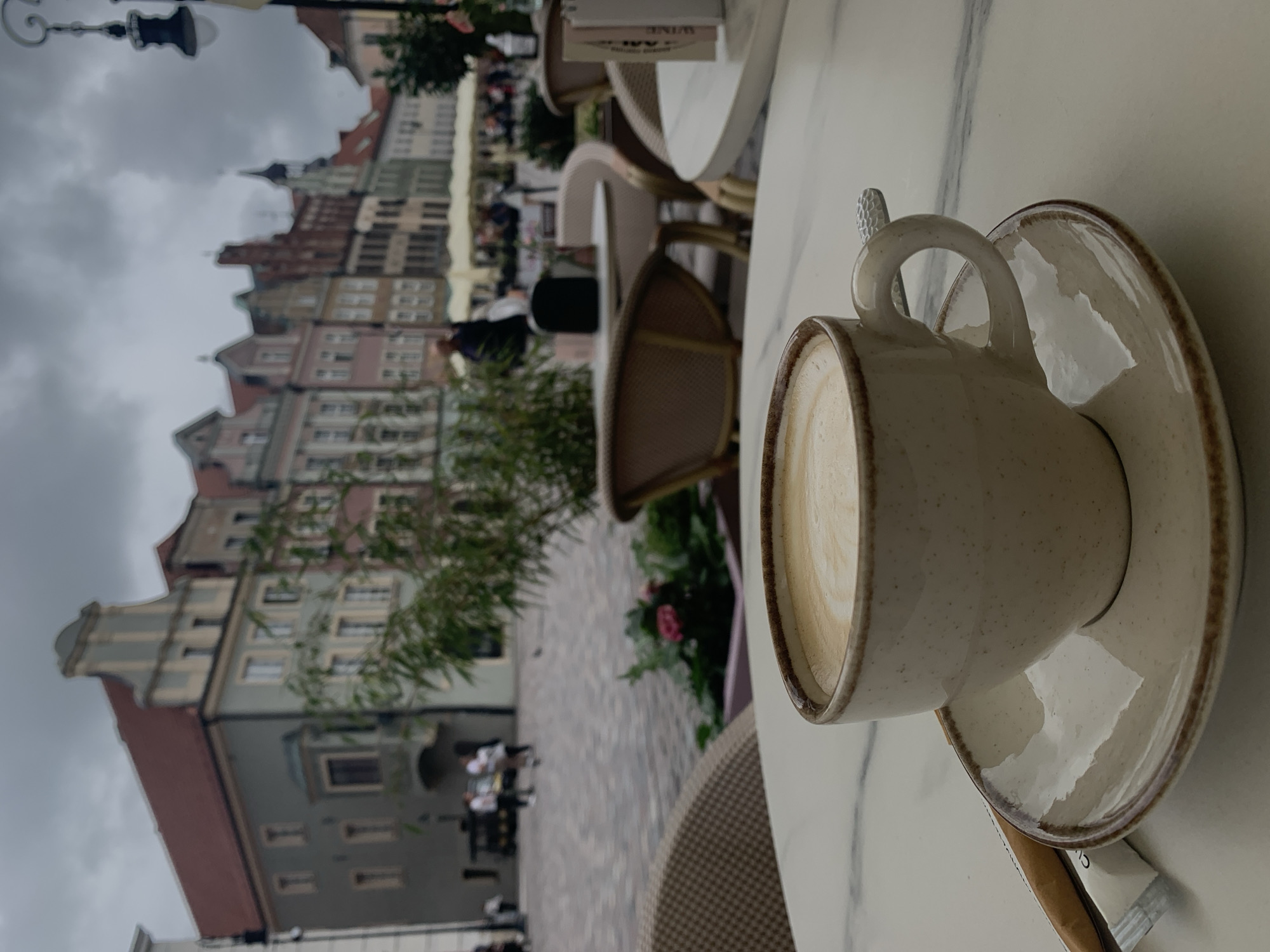 A close-up shot shows a cup of coffee with latte art on a marble tabletop. The coffee cup and saucer are speckled and have a brown rim. To the left of the cup is a sugar packet and a stir stick. In the background, the scene is blurry, depicting a European city square with colorful buildings and outdoor seating at a cafe. The architecture suggests a historic European city, consistent with the location being the Old Market Square in Poznan, Poland. The sky is overcast. A few people are visible in the background, walking through the square.