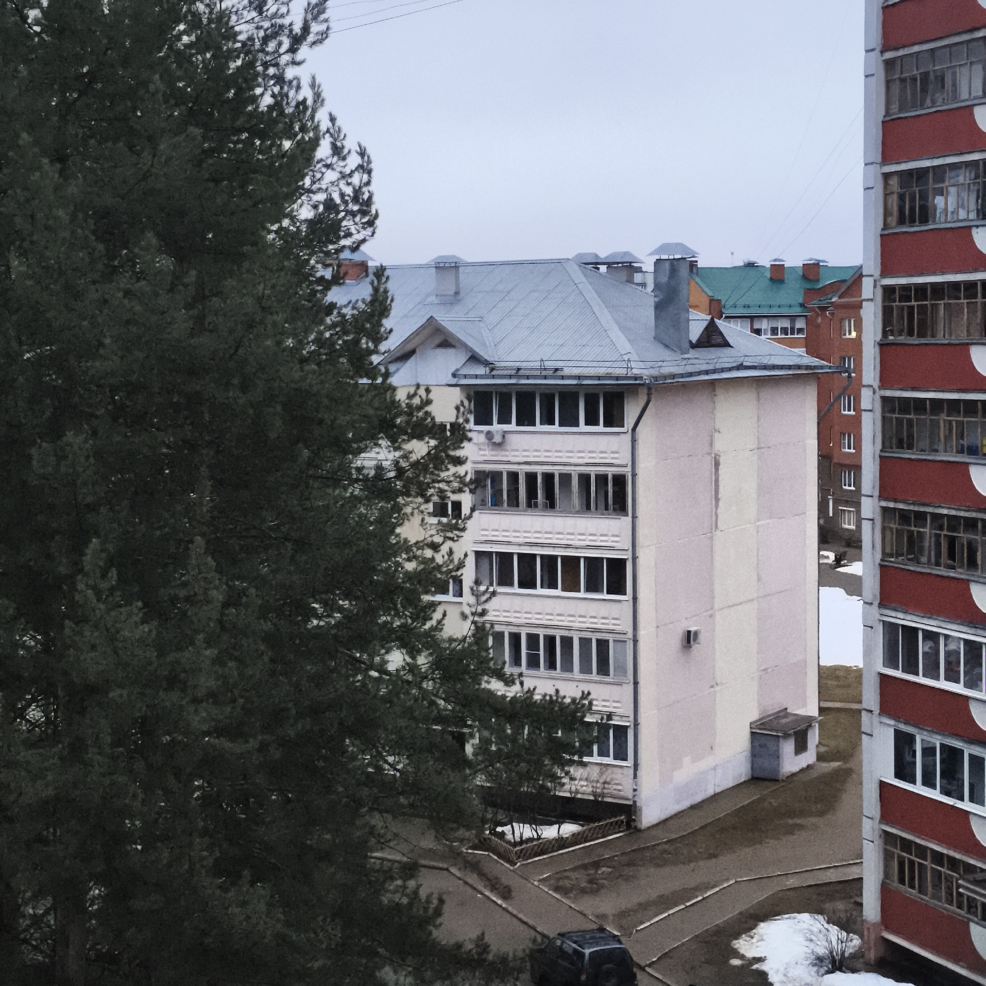 The image shows an urban residential scene. In the foreground to the left is a large evergreen tree. To the right of the tree is a light-colored, four-story residential building with a metal roof and several chimneys. A red and white building is on the right side of the frame, partially obscuring the view. A black SUV is parked on the street below. Patches of snow are visible on the ground. In the background are additional buildings with varying roof colors. The sky is overcast.