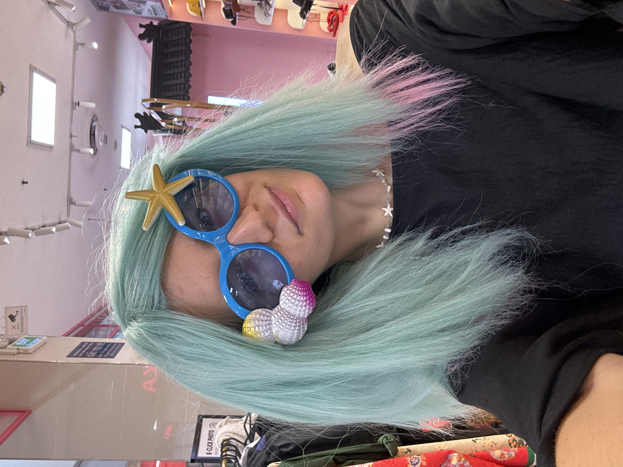 The image shows a person with light blue and pink hair wearing novelty sunglasses with a starfish on one side and a three-sphere decoration on the other. They are wearing a black shirt and a star-shaped charm necklace. The background suggests an indoor retail environment with pink walls and overhead lighting, as well as a counter and various items for sale. A sign that includes the words "K-CLICK PHOTO" is visible in the lower-left corner. Based on this information, the location is possibly in the city of Phú Thủy, Vietnam.
