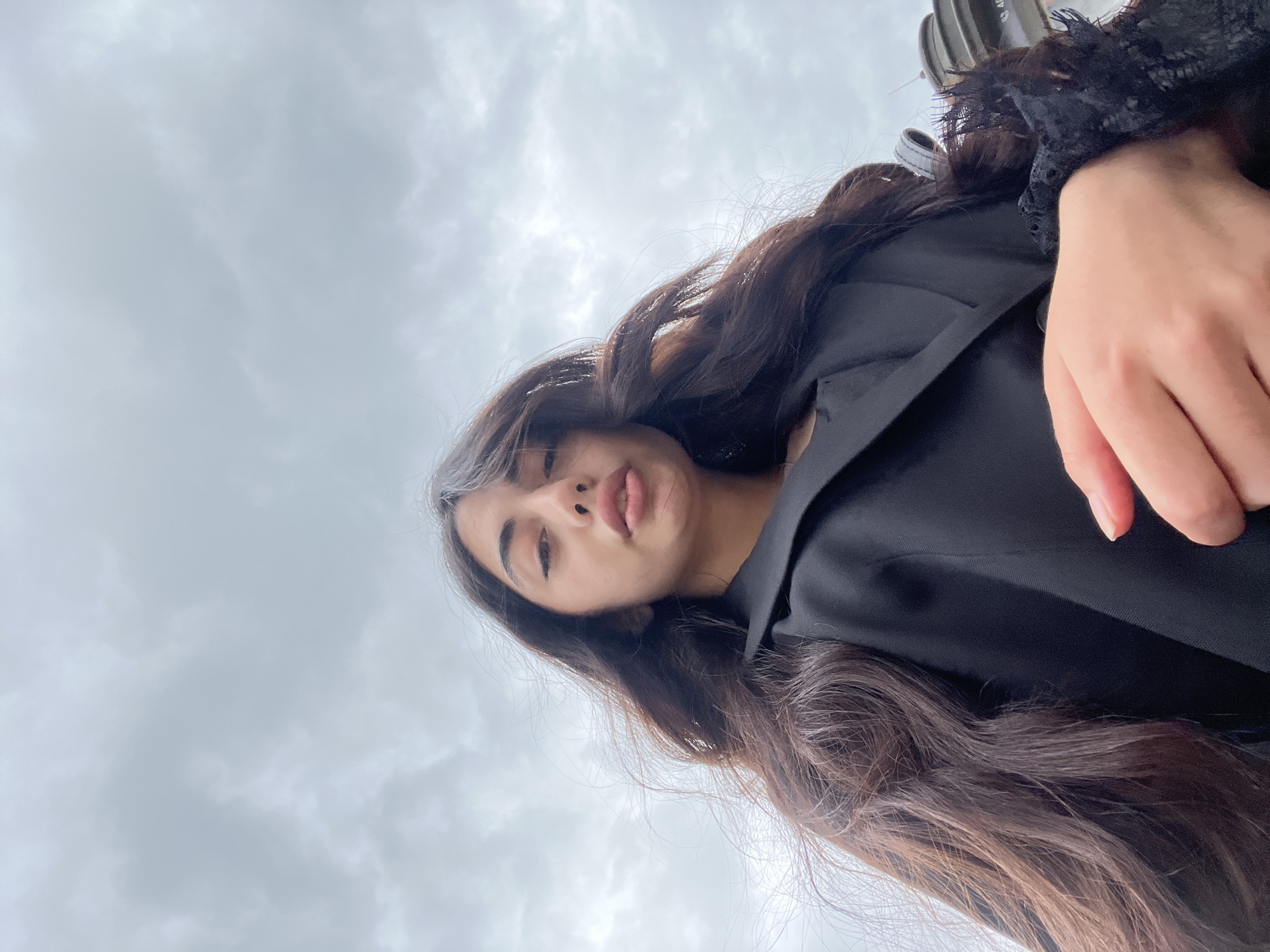 A low-angle, close-up shot shows a young woman with long, dark, wavy hair against a cloudy sky. She is wearing a black blazer with lace detailing on the cuffs. Her face is turned towards the camera, and she is looking directly at the viewer. Her mouth is slightly open, and her lips are plump and pink. Her right hand is visible in the foreground, with her fingers slightly curled. The background is dominated by a grey, overcast sky with visible cloud formations. The image appears to be a selfie taken outdoors. The presence of a distinctive circular object, possibly a monument or sculpture, is visible in the upper right corner. This object, along with the architecture visible behind it, suggests the location is the Republic Square in Pilsen, Czechia.
