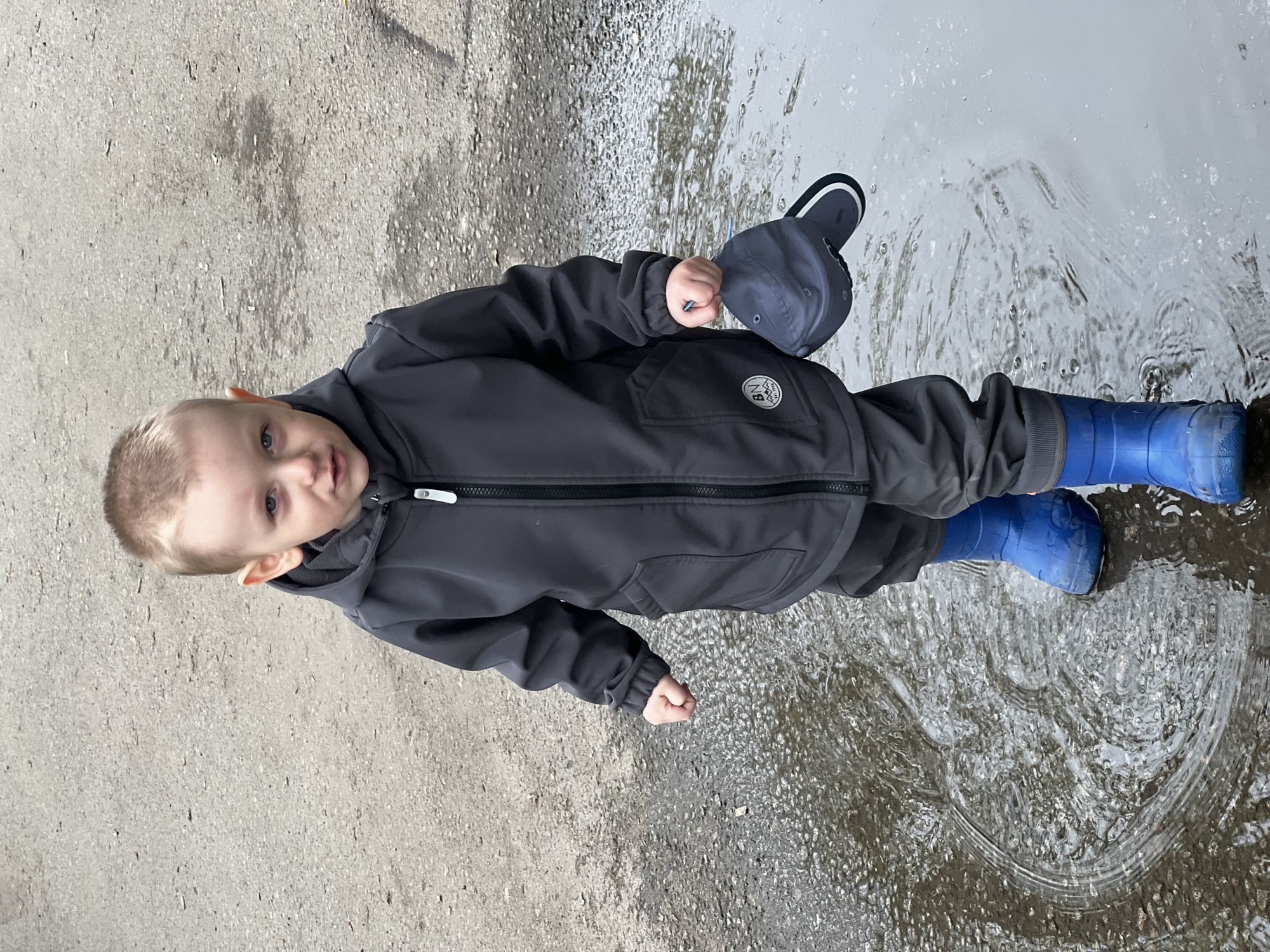 A young child, likely a boy, stands partially in a large puddle on a paved surface in Mytishchi, Russia. The child has light-colored hair and blue eyes, and looks directly at the camera with a calm expression.
He is dressed in a dark gray or black hooded jacket with dark pants, and bright blue rubber boots that are submerged in the water, creating ripples. The jacket features a small, circular white emblem on the left chest area, though the specific details of its design or text are not clearly legible. In his right hand, the child holds a dark blue baseball cap.
The setting appears to be outdoors on a daytime, possibly overcast day, given the even lighting and the presence of the puddle. The ground is a mix of dry, gray asphalt or concrete and a significant area of standing water, indicating wet conditions or recent rain. The activity shows the child engaging with the puddle, characteristic of childhood exploration.
