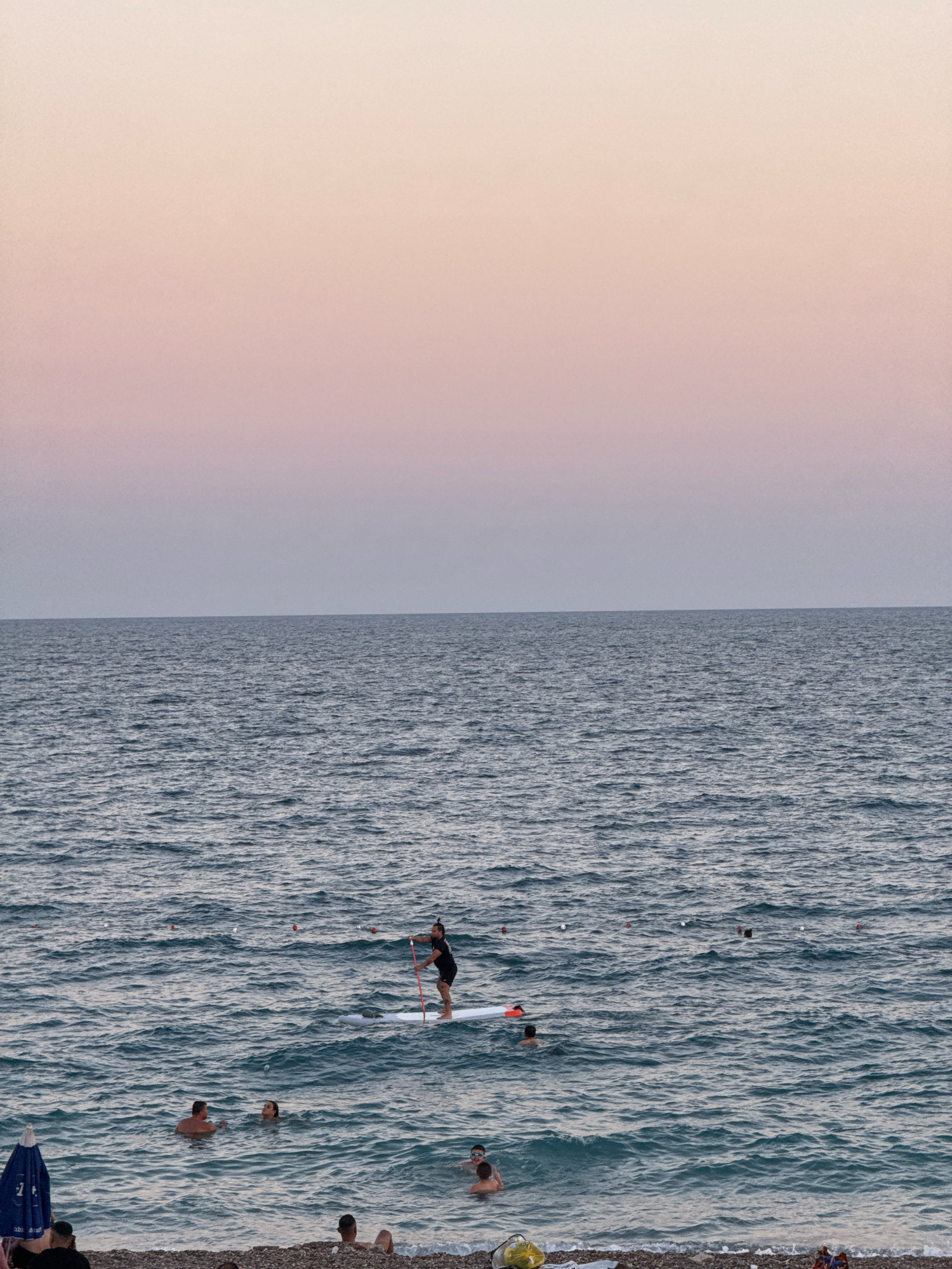 The image captures a serene scene at Konyaaltı Beach in Antalya, Turkey, during what appears to be either sunrise or sunset, given the soft pastel gradient of the sky transitioning from pale pink to light grey. The vast expanse of the Mediterranean Sea dominates the frame, its surface rippled by gentle waves.
In the middle ground, a person is paddleboarding on a white board, holding a paddle with a red shaft. The individual is wearing dark clothing. Several other people are visible in the water, some swimming and others wading closer to the shore. One group of two appears to be conversing.
The foreground features the pebbled beach, with a partially visible blue beach umbrella on the left. A few individuals are seated or lying on the beach, enjoying the atmosphere. Small buoys or markers are scattered across the water's surface, suggesting a designated swimming or activity area. The overall impression is one of a peaceful evening or morning by the sea.