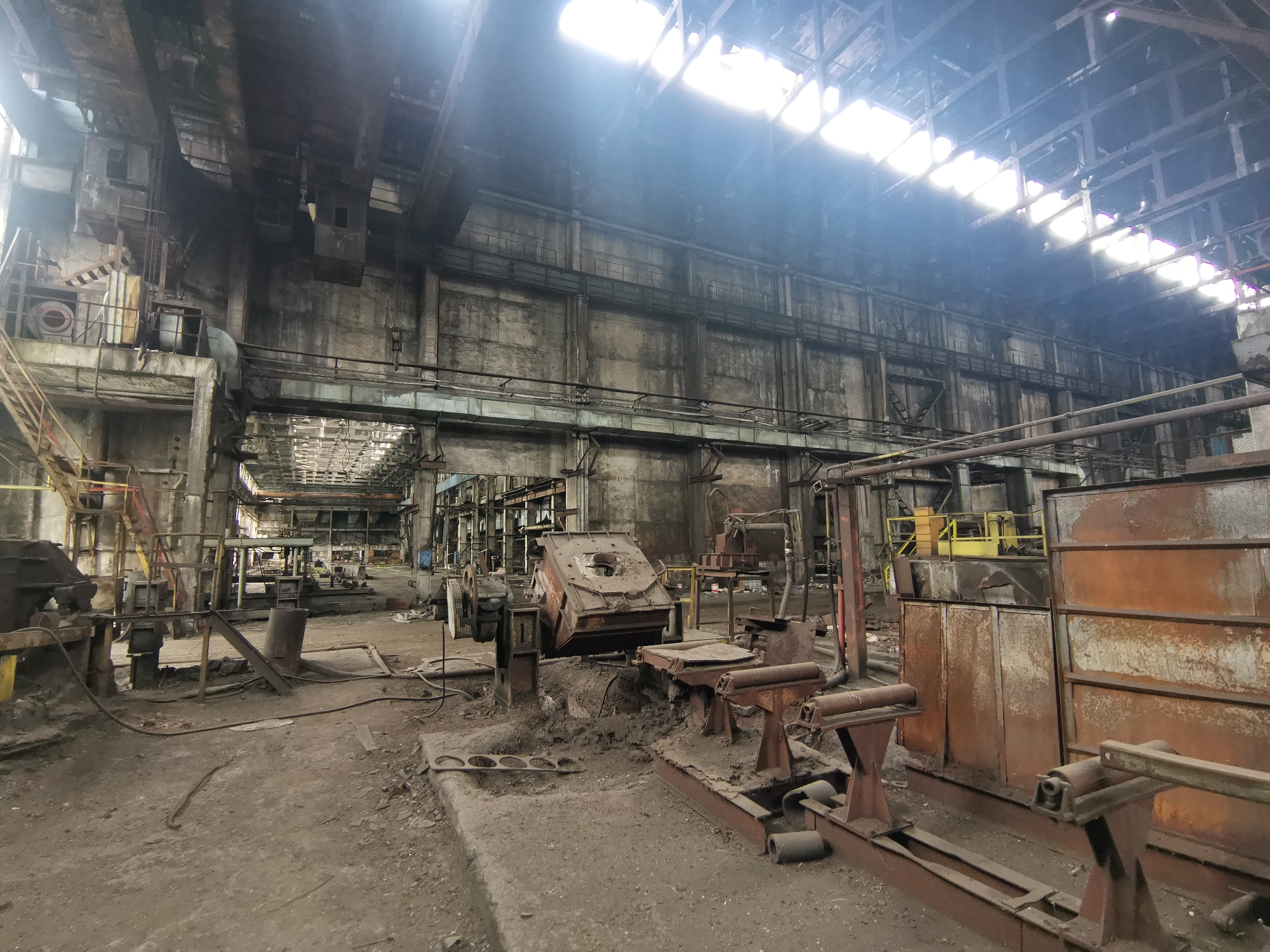 The image shows the interior of an abandoned industrial building. The scene appears to be a derelict factory floor with a high ceiling, likely a foundry or metalworking plant, as evidenced by the presence of heavy machinery, casting equipment, and metal structures. The walls are stained and dilapidated, indicating significant neglect and disuse. Natural light filters through broken skylights, illuminating the dust-covered surfaces and scattered debris. The machinery is rusted and appears to be non-operational. No people are visible in the frame. There is no visual evidence to confirm the location as Zhivkovo, Bulgaria. The scene depicts a state of abandonment and industrial decay.