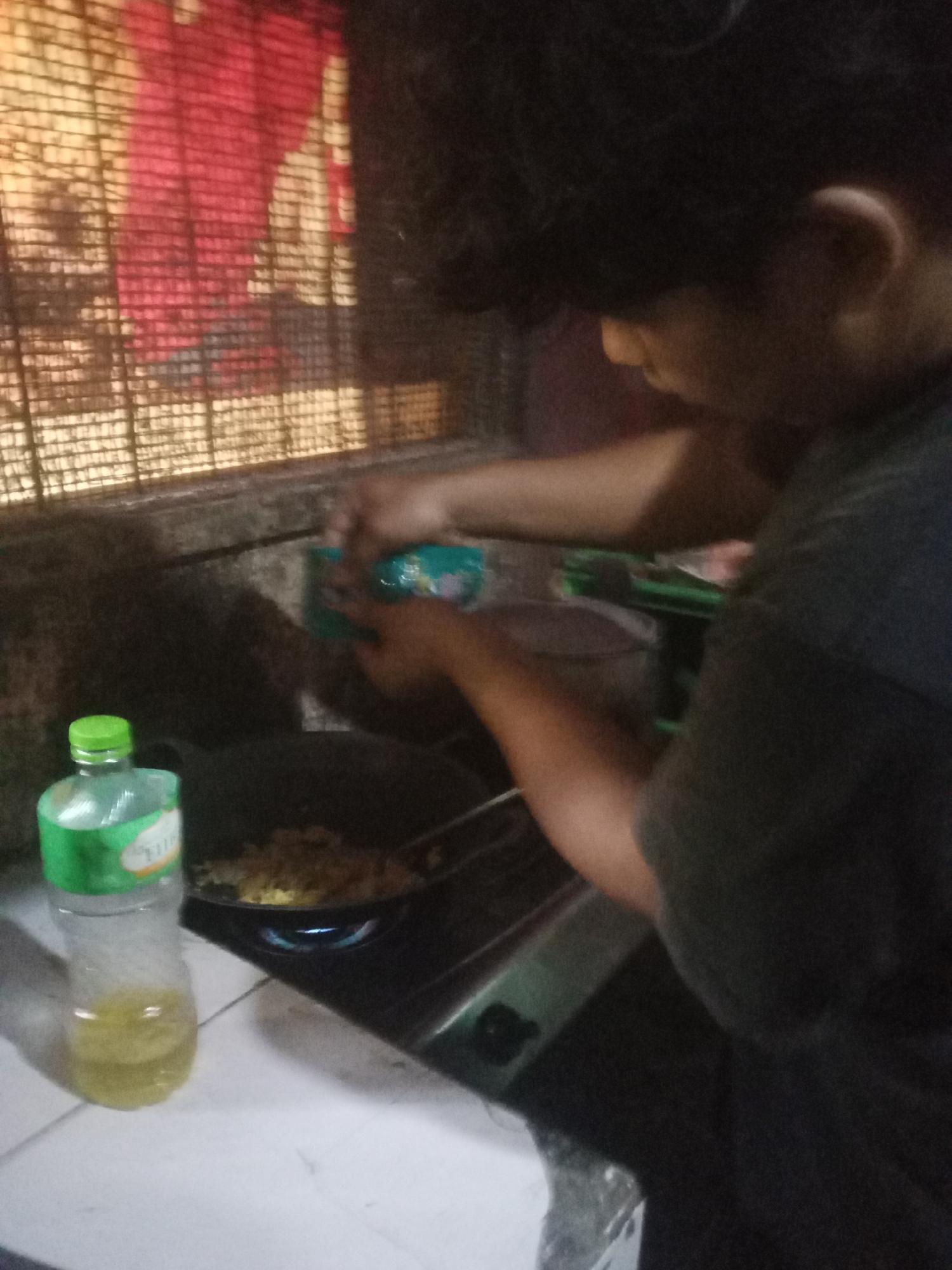 The image shows a person cooking in a kitchen, likely in Cintaratu, Indonesia. A bottle of cooking oil with a green cap is visible in the foreground, next to a gas stove where food is being prepared in a pan. The background features a patterned curtain or screen. The person's hands are engaged in the cooking process, stirring the contents of the pan. The lighting is dim, creating a slightly grainy appearance.