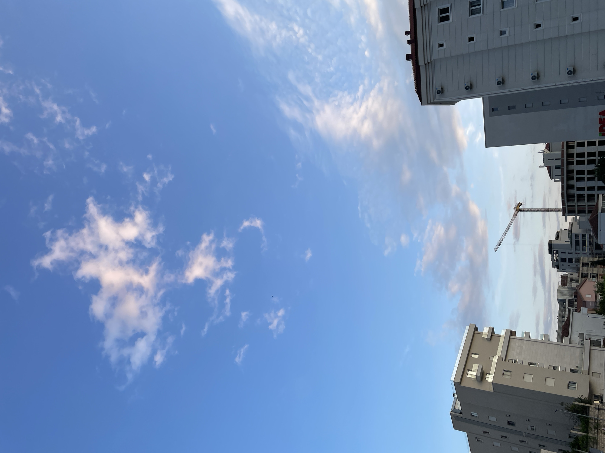 The image depicts a low-angle view of the sky and surrounding buildings in Boreti, Montenegro. The sky is primarily blue with scattered white clouds. Buildings are visible on the right and bottom edges of the frame. One building appears to have a sign that says "IDEA". A construction crane is visible amidst the buildings. No people are discernible in the image.