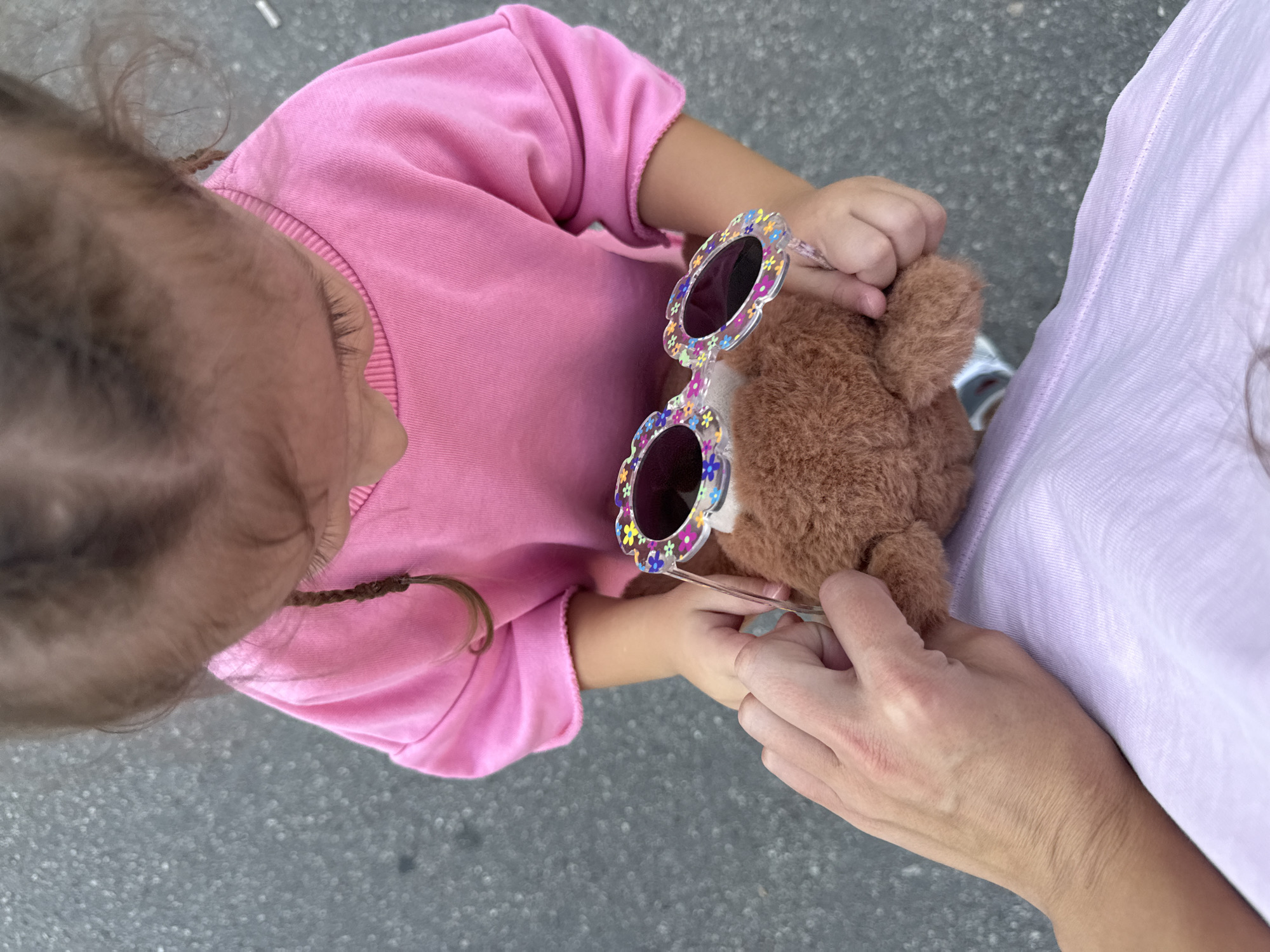 A young child wearing a pink sweatshirt and a person's hand are in the foreground of the image. The child holds a small brown teddy bear and is wearing flower-shaped sunglasses. The person's hand is holding the child's hand. They are standing on a grey, asphalt-like surface, possibly a sidewalk or road. The background is blurred and shows a grey, textured ground. The image is taken from a high-angle perspective. The location is not specified in the image and cannot be determined.