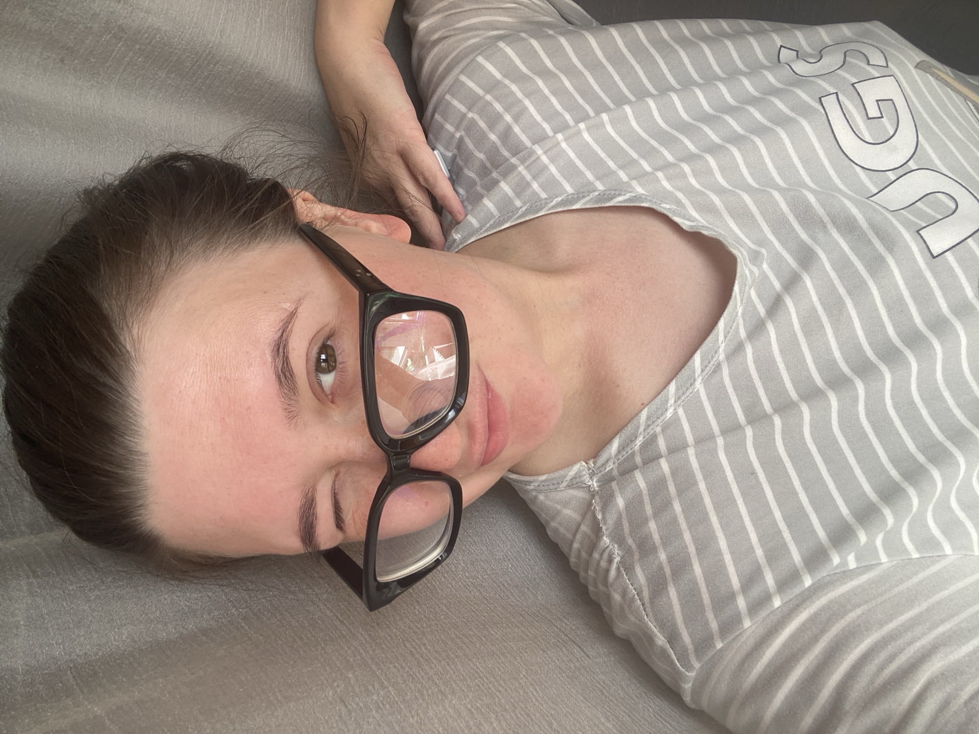 A young woman is depicted lying down on a textured grey surface, likely a couch or bed, in an indoor setting. She has dark hair, styled away from her face, and is wearing black-framed glasses. Her head is tilted, and she looks directly towards the viewer with her right eye open and her left eye partially closed, giving the impression of a wink or blink. A slight smile plays on her lips. She is wearing a light grey top with thin white vertical stripes. The brand name "UGG" is visible in white letters on the fabric of her shirt, positioned towards the top right portion of the image. Her right arm is visible, with her hand resting near her ear. The lighting is soft and even, without strong shadows, indicating a relaxed atmosphere. The context provided for the location is Chaikovsky, Russia.