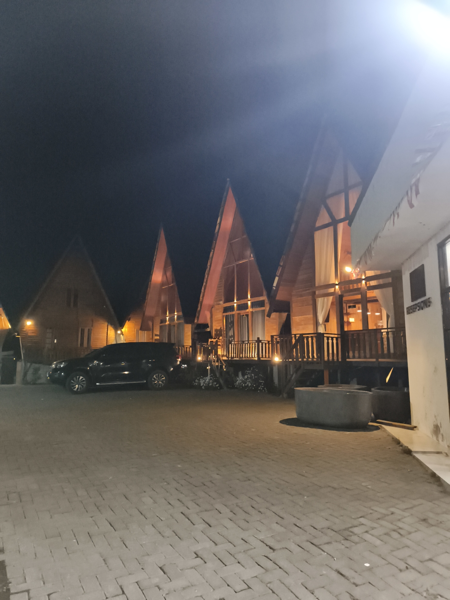 In the city of Dieng Kulon, Indonesia, a black SUV is parked on a brick-paved surface at night. The vehicle is positioned to the left of several A-frame wooden cabins. These cabins feature large glass windows and wooden balconies, with interior lights illuminating the spaces. One of the cabins has a sign reading "RESEPSIONIS" on its white exterior wall. In the foreground, two large, grey concrete planters are visible on the paved area. The scene is illuminated by artificial lighting, with a bright light source creating a flare in the upper right portion of the image. The background is dark, indicating it is nighttime.