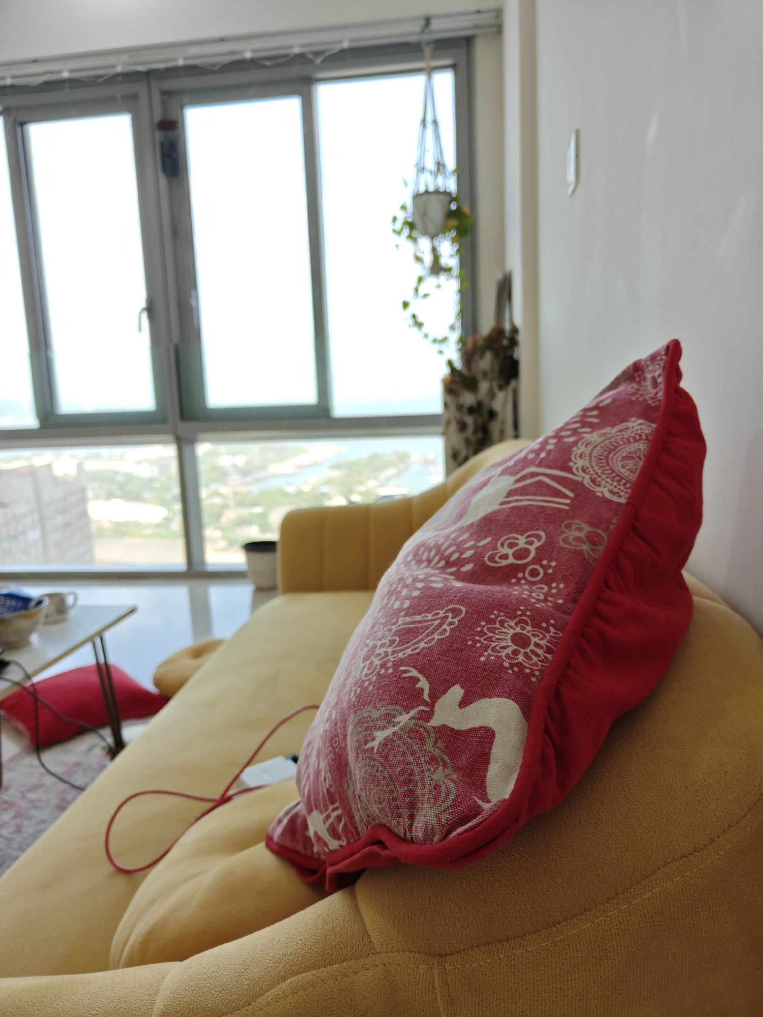 A medium shot focuses on a red and white decorative pillow placed on a yellow couch. The pillow is detailed with a complex pattern and a red ruffle trim. A thin red cable snakes across the couch towards an unseen device. In the background, large windows reveal a partial view of what appears to be a city, suggesting an urban location. The presence of water bodies, seen through the windows, along with building clusters, indicates a coastal metropolitan area, suggestive of Mumbai, India. A plant hangs near the window, adding a touch of domesticity to the scene.