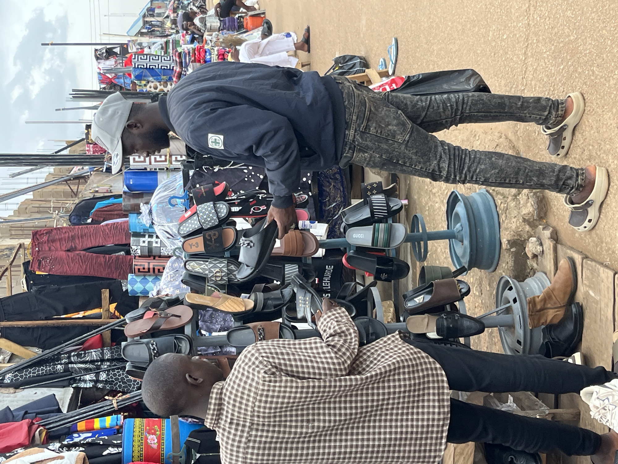 This image captures a scene at an outdoor market in Bukuru, Nigeria. Two men are the primary subjects, positioned amidst stalls laden with footwear and clothing. On the left, a man wearing a brown plaid long-sleeved shirt and black pants is examining a display of sandals. He holds one of the sandals in his hands. To his right, a man in a navy blue jacket, dark distressed jeans, and a grey baseball cap is looking towards the sandal display, with one hand extended towards a black sandal.
The environment is characterized by numerous stalls featuring a variety of items, predominantly shoes and garments. Several racks are filled with different types of sandals, including slides and flip-flops, some of which appear to be designer brands like Gucci. Other clothing items, such as patterned pants and shirts, are also visible hanging or displayed. The ground is a dirt or sandy surface, typical of an outdoor market. In the background, other market-goers can be seen, suggesting a busy commercial area. The sky is overcast, indicating a cloudy day. The overall atmosphere is that of a lively marketplace.