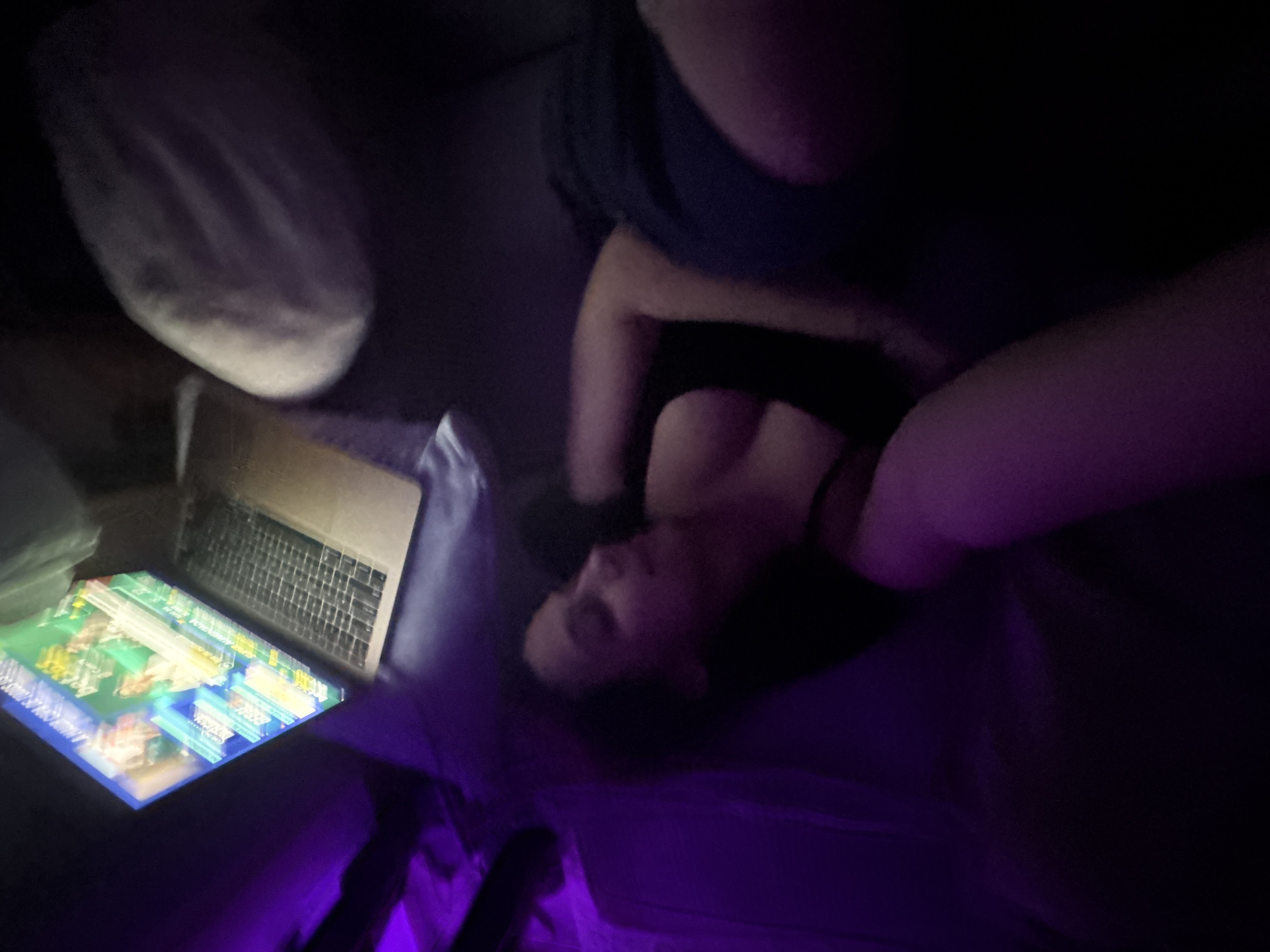 The image depicts a dimly lit indoor scene, possibly a living room, where a woman is lying down. A laptop is open in front of her, displaying a colorful interface, likely a website or application. The ambient lighting is predominantly purple, casting an unusual hue over the environment. The woman is wearing a dark top and dark shorts. Her body language suggests she is relaxed or resting. The overall impression is that of a private, intimate setting, possibly late at night, where the woman is engaged with her laptop. There is no identifiable information that can pinpoint the location to Mönchengladbach, Germany.