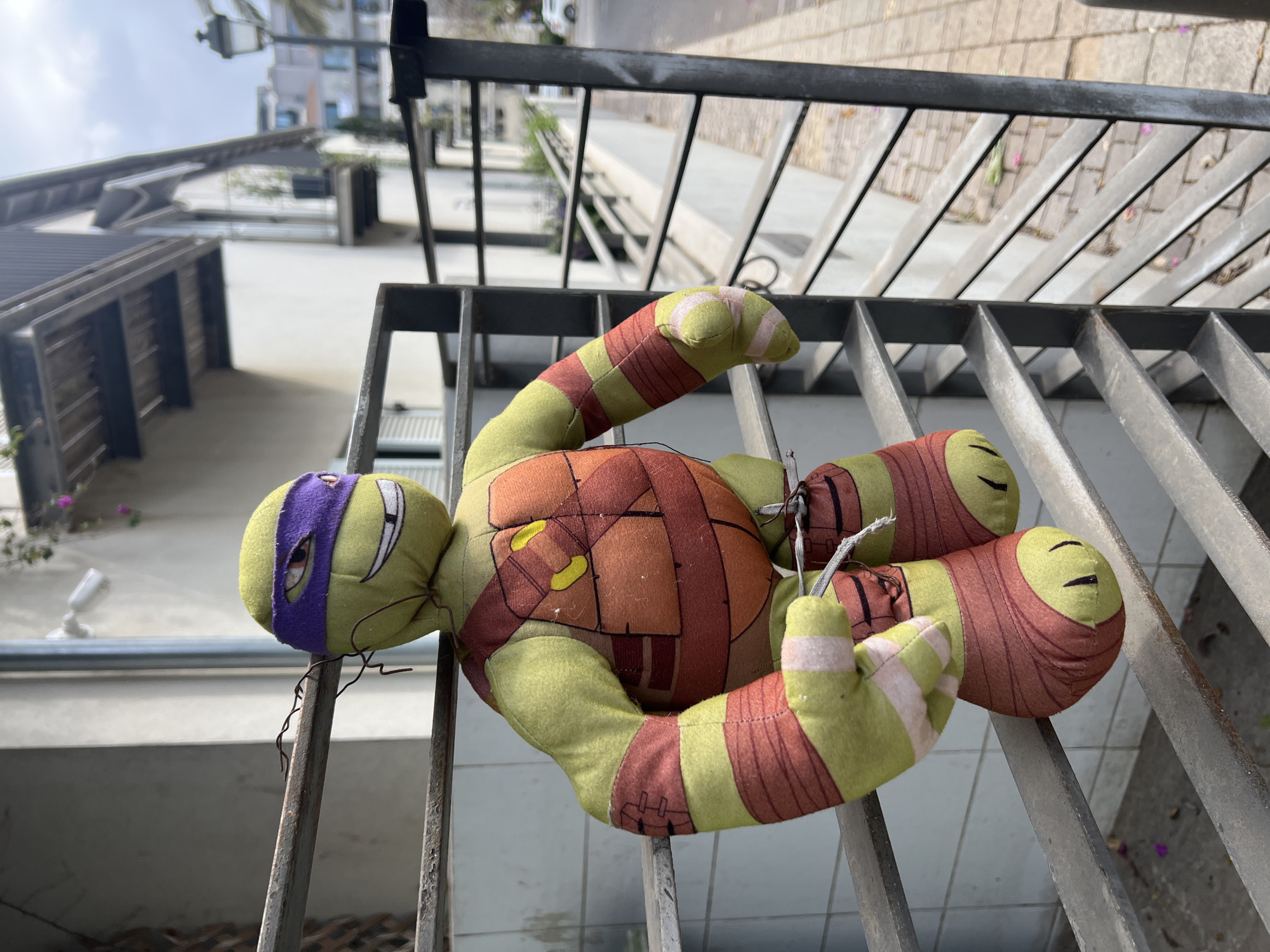 A plush Teenage Mutant Ninja Turtle toy, specifically Donatello wearing a purple mask, lies entangled within the metal bars of a railing in Tel-Aviv, Israel. The toy appears worn. The background reveals a cityscape, suggesting an urban environment. The toy is positioned on the metal railing, with parts of its body and limbs caught between the bars. The railing overlooks a patio area paved with concrete. In the background, buildings with modern architectural features can be seen. The scene is outdoors during the day.