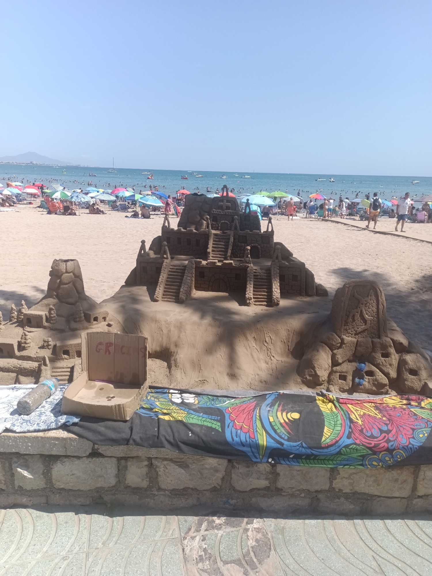 On a sunny day at the beach in Peníscola, Spain, a large, elaborate sandcastle depicting a fortress-like structure with multiple levels, stairs, and towers dominates the foreground. The sandcastle is intricately designed and appears to be inspired by ancient architecture. In front of the sandcastle, a cardboard box with the word "GRACIAS" written on it, possibly for donations, sits atop a colorful patterned cloth. A plastic water bottle is also visible. The background of the image shows a crowded beach with numerous beach umbrellas, people sunbathing and swimming in the blue ocean, and boats on the water. The sky is clear and blue.