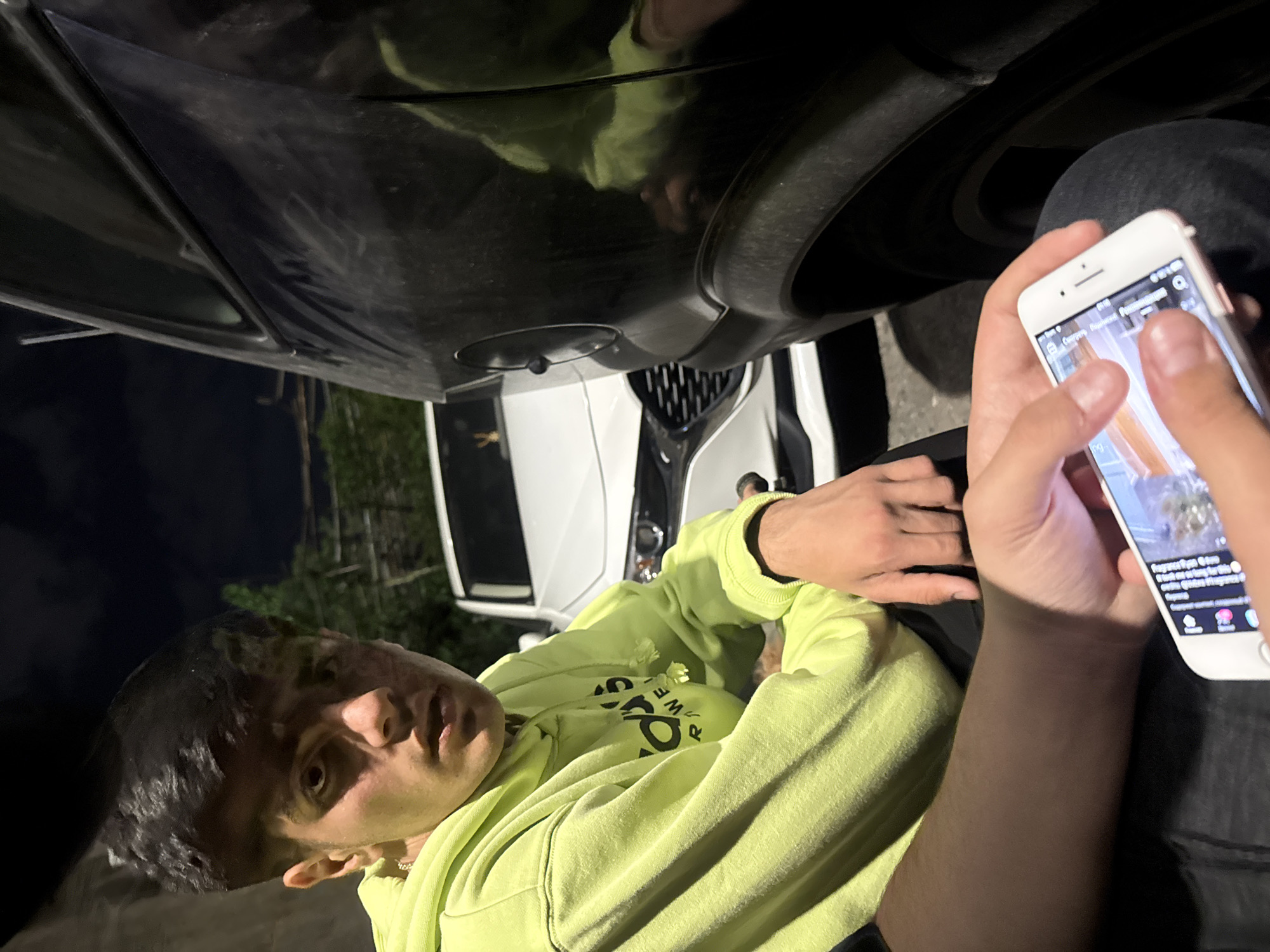 The image shows a young man lying on his back, partially under a car, looking at the camera. He is wearing a neon yellow hoodie. He is holding a white smartphone in his right hand and looking at the screen. The car appears to be parked on a street at night, with trees visible in the background. Another, white, car is also visible. Given the request, the exact location is Yerevan, Armenia.