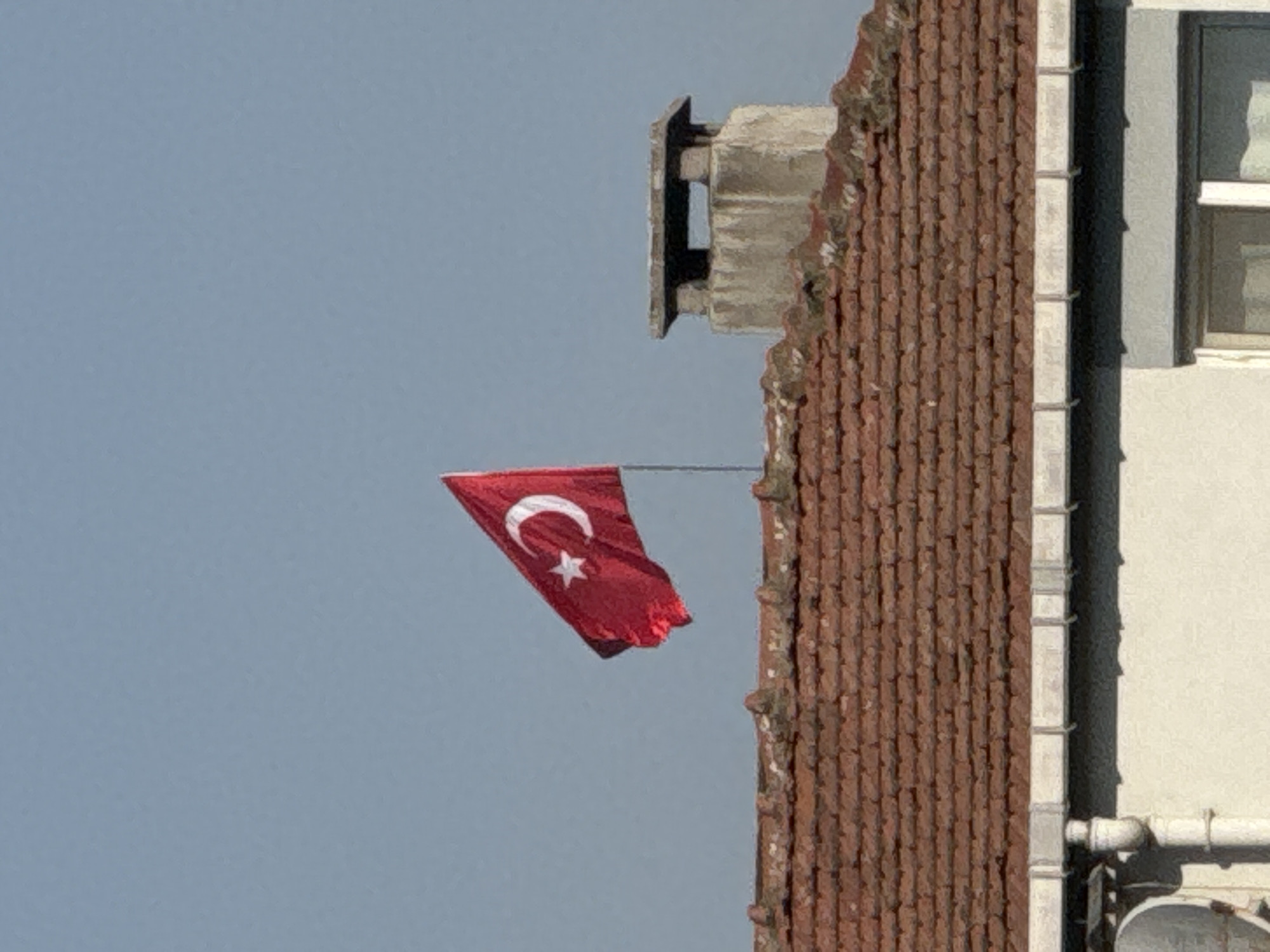 A Turkish flag flies from a flagpole on a rooftop in Gungoren, Turkey. The flag is red with a white crescent moon and star. The roof is covered in red tiles. To the right of the flag is a stone chimney. In the foreground, part of a white building with a gutter is visible. The sky is a clear blue.