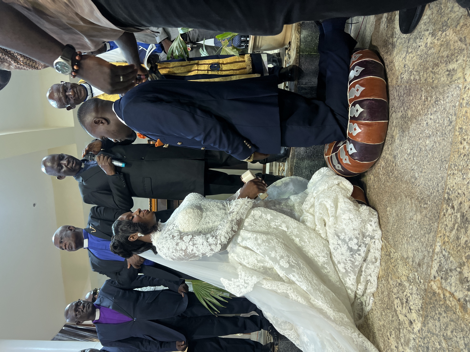 The image depicts a wedding ceremony in Kabong, Nigeria. The bride, wearing an elaborate white lace wedding gown and veil, is kneeling on a decorative cushion in the foreground. The groom, in a dark blue suit with gold buttons, is also kneeling and appears to be proposing or exchanging vows. Several men in formal attire, including some wearing clerical collars and robes, stand behind the couple, suggesting they are officiating the ceremony or are guests of honor. The background shows an indoor setting with stone flooring and what appears to be a church or formal venue. The presence of palm fronds suggests a tropical or sub-tropical location.