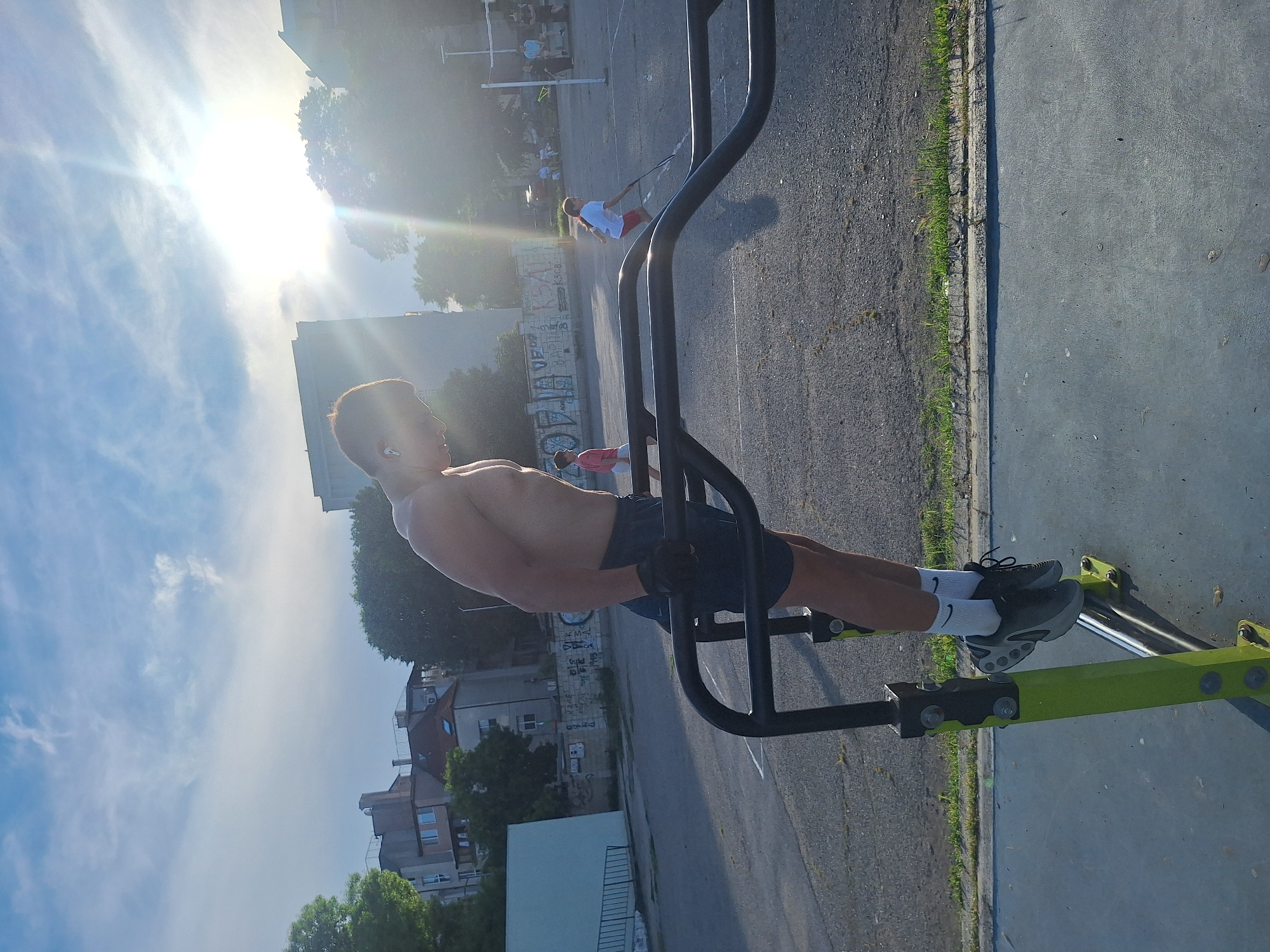 A shirtless young man is performing dips on parallel bars in an outdoor urban park in Varna, Bulgaria. He is wearing dark shorts, black gloves, white socks with black and grey athletic shoes. The sun is shining brightly in the sky, creating a lens flare effect. In the background, other people are visible, including a child playing with a racket and a net, suggesting a sports or recreation area. Buildings and trees form the backdrop, typical of a city environment. The ground is paved with asphalt.