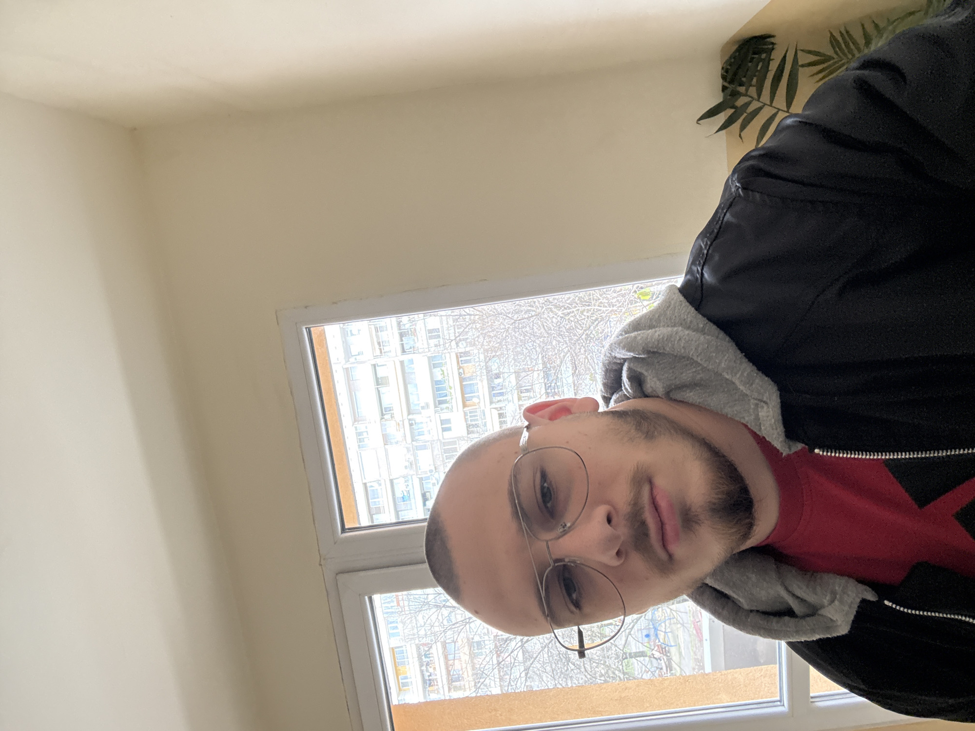 A man with a bald head and beard, wearing round-framed glasses, a red shirt, and a black jacket with a gray hoodie, is positioned in front of a window. The window offers a view of a building exterior with numerous windows. The scene appears to be indoors, potentially within an apartment. The location is likely Stara Zagora, Bulgaria.
