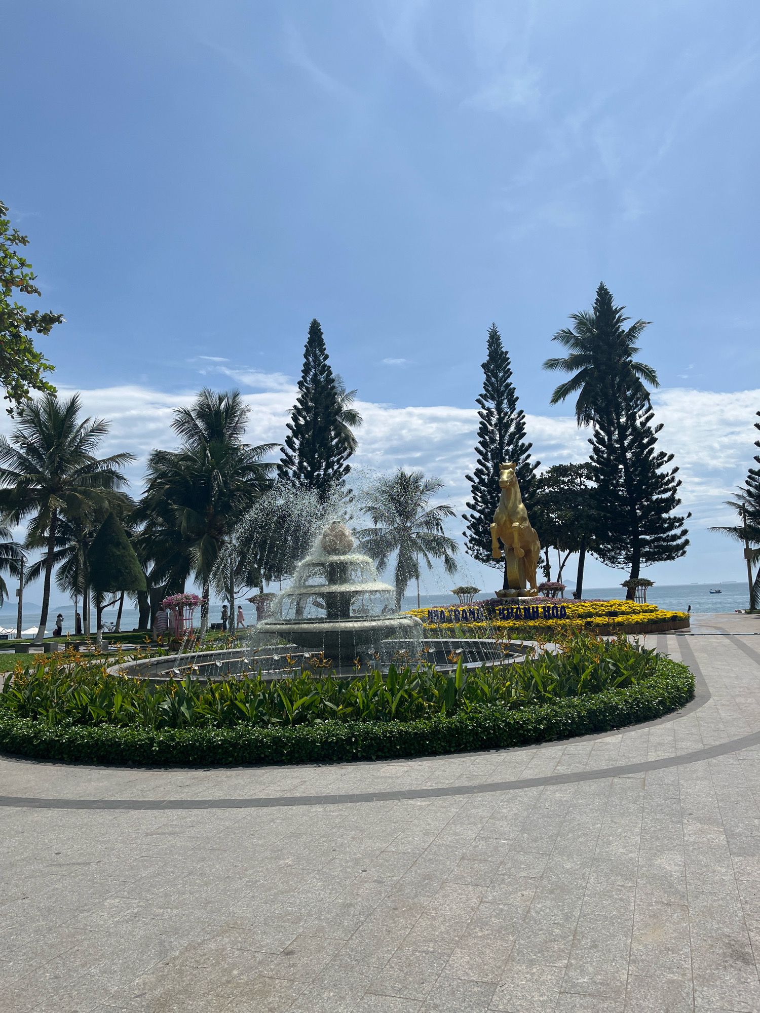 This image captures a public plaza in Nha Trang, Vietnam, featuring a central fountain and a prominent golden horse statue. The scene is set against a backdrop of palm trees and tall pine trees, with the ocean visible in the distance. The fountain, located in the foreground, is actively spraying water, creating a dynamic visual element. Behind the fountain, the golden horse statue stands on a pedestal adorned with yellow flowers, forming the centerpiece of the plaza. Several people are present, appearing to be casually walking or enjoying the outdoor space. The ground is paved with stone tiles, and manicured greenery surrounds the fountain and flower beds. The sky is blue with scattered clouds, suggesting pleasant weather. The overall impression is one of a well-maintained urban park or promenade, likely a popular spot for locals and tourists. The inscription on the flower bed behind the horse statue appears to read "NHA TRANG KHANH HOA", indicating the specific location.
