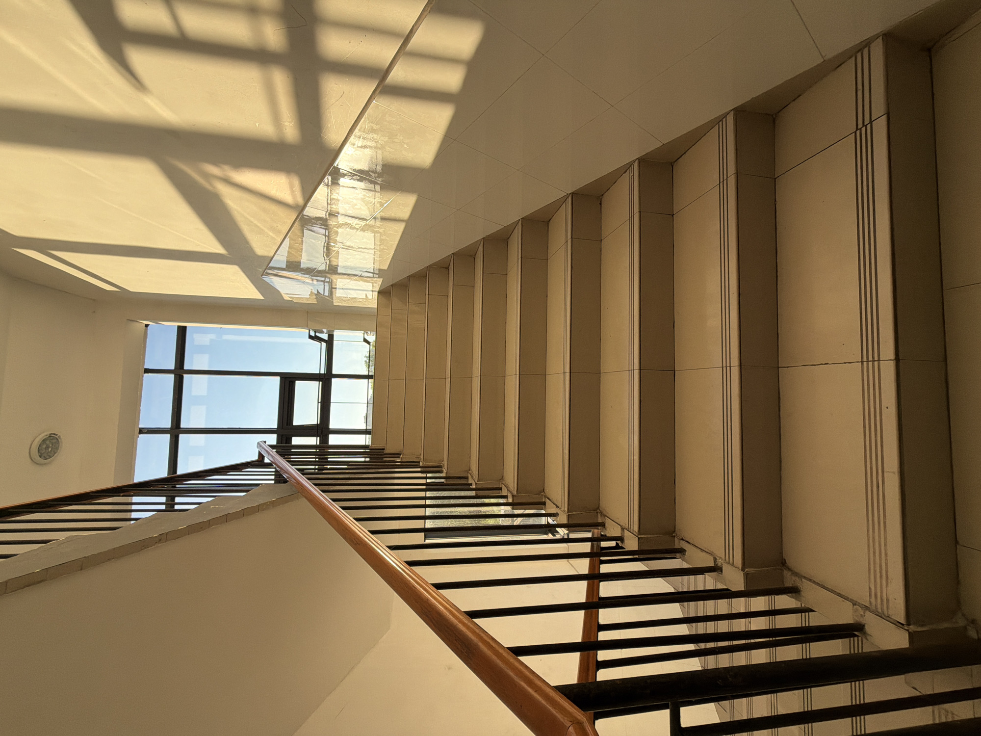 The image shows a flight of stairs in what appears to be an indoor public building, possibly an apartment complex or office building, located in the city of Jiangjin, China. The stairs are made of light-colored tiles with darker accent lines on the edges of each step. A dark metal railing with a wooden handrail runs along the left side of the staircase, and a similar railing is visible on the right side, though partially obscured. Natural light streams in from a large window at the top of the stairs, casting distinct shadows of window panes and a railing onto the wall on the right. The window overlooks a bright sky and some distant greenery. The overall scene is clean and well-lit, with no people or animals present. The architectural style suggests a modern construction.