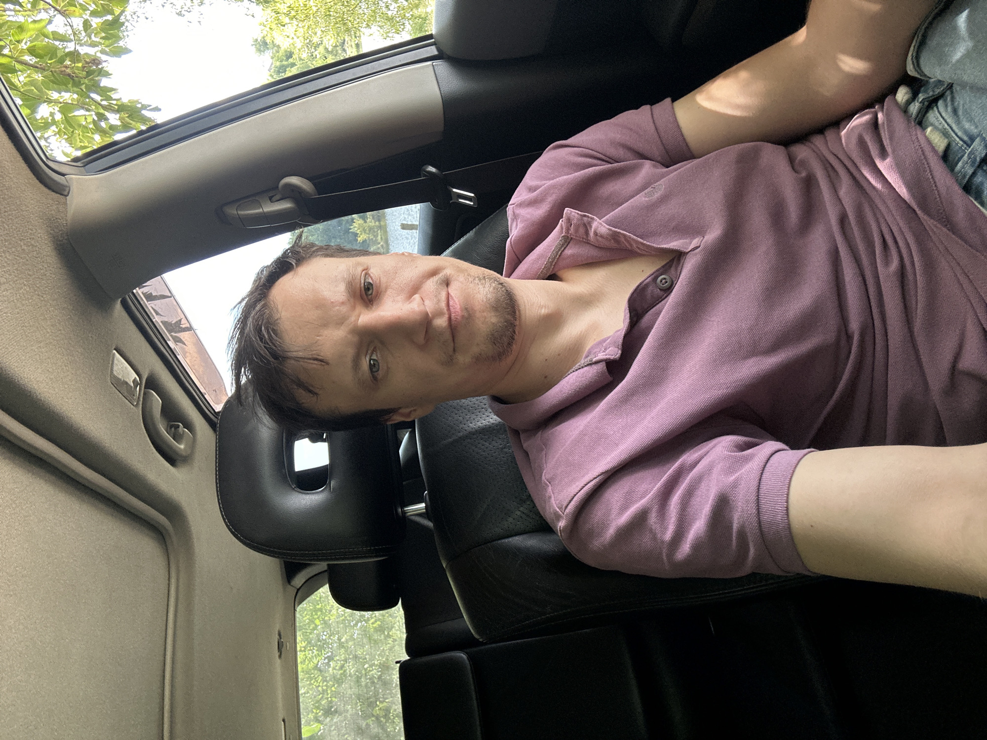 A man is sitting in the backseat of a car, which appears to be an SUV, judging by the headrests and the overall interior. He is looking directly at the camera and smiling slightly. He is wearing a purple polo shirt and blue jeans. The car's interior is dark leather, and there is a sunroof open above. Through the windows, we can see trees and water, suggesting the car is parked in a natural setting, possibly near a lake or river. The image is taken from a close-up perspective, focusing on the man's face and upper body. There is no explicit indication of the location being Makiv, Ukraine, as the image content does not provide such specific information.