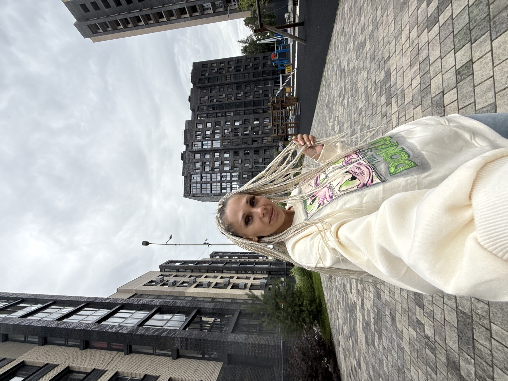 A woman with blonde braided hair is standing in front of apartment buildings in Rusanovo, Russia. She is wearing a cream-colored sweatshirt with a cartoon graphic on the front. The background consists of tall, modern apartment buildings with dark facades and numerous windows. There is a playground in the middle ground. The ground is paved with gray and beige rectangular tiles. The sky is overcast.