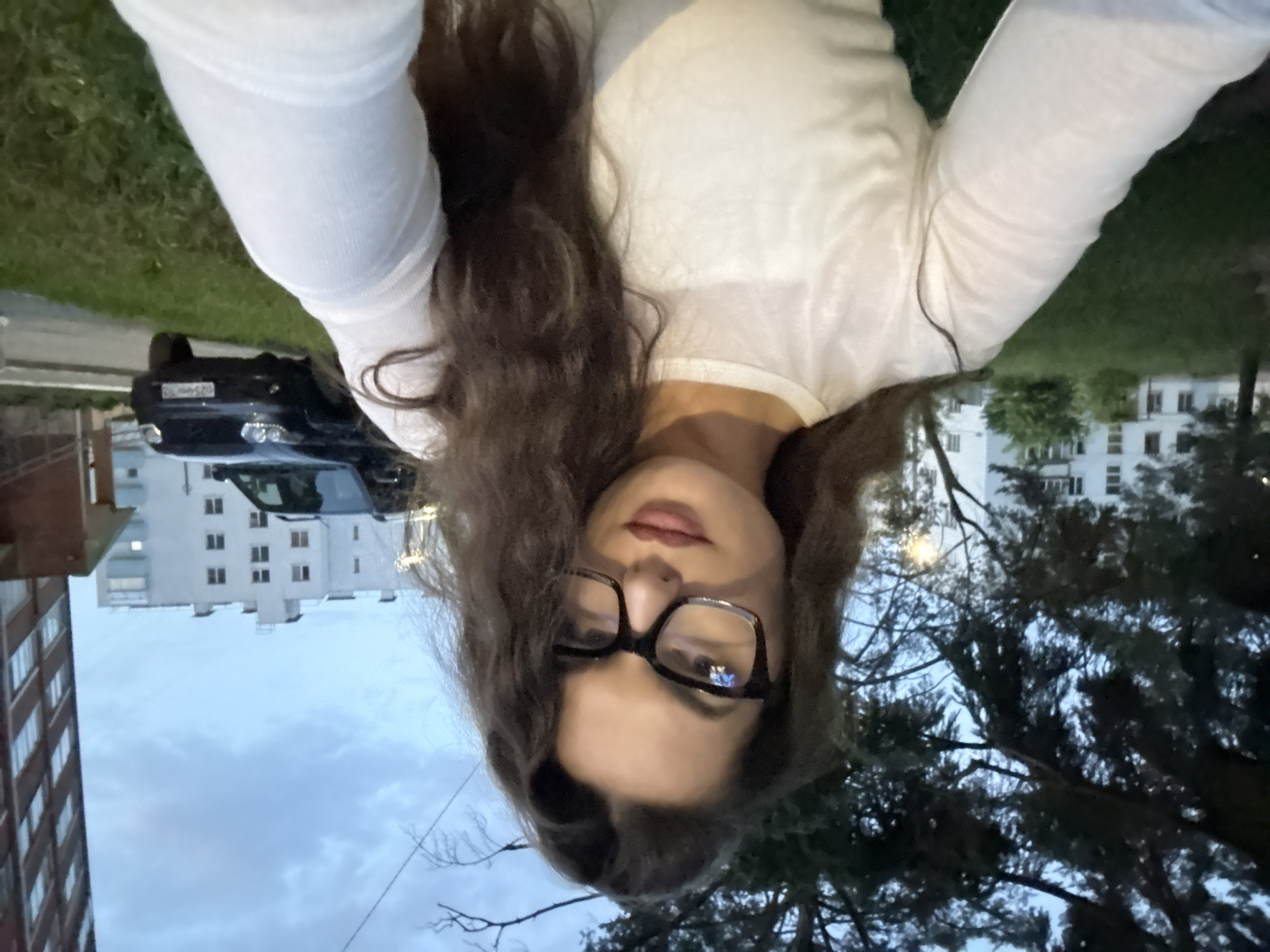 A medium-shot selfie shows a young woman with long, dark, wavy hair and a white long-sleeved shirt. She is wearing black-rimmed glasses and has on makeup. The background suggests an urban environment, possibly in the city of Chaikovsky, Russia. To the left of the woman, there are trees and a glimpse of a white apartment building. To her right, a dark-colored SUV is parked on a grassy area in front of a brick building, with another white apartment building visible further in the background. The sky is overcast with clouds.