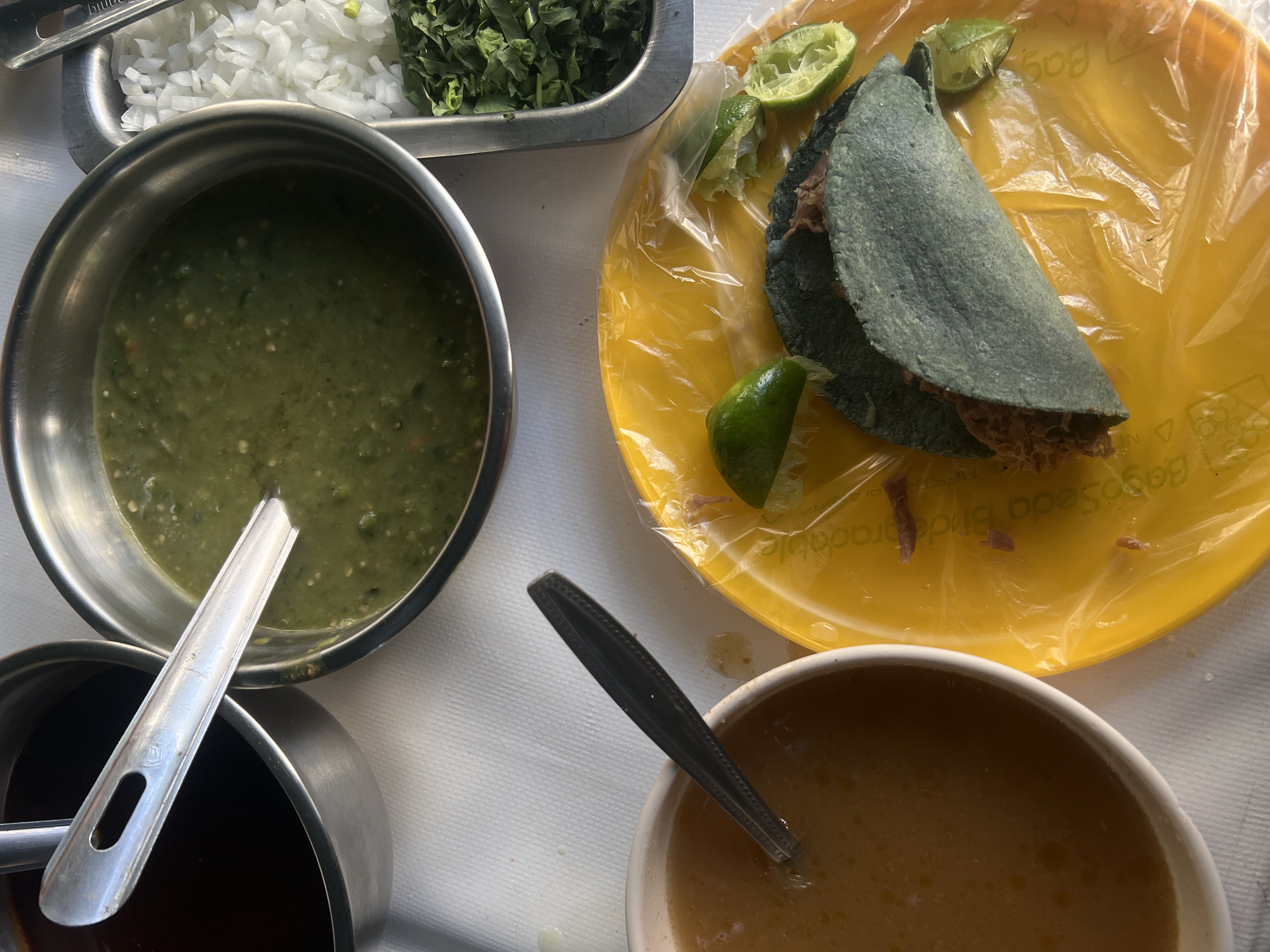 The image depicts a meal of tacos served in Mexico City, Mexico. A blue corn tortilla taco is placed on a yellow plate with lime wedges. Condiments are presented in metal and ceramic containers, including salsa verde, salsa roja, and a light brown consommé. Additional toppings of chopped onions and cilantro are in a metal tray. Spoons are in the salsa and consommé. The meal is set on a white surface.