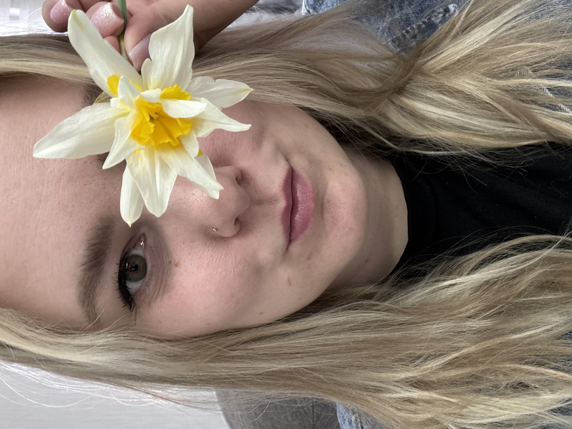 A fair-skinned woman with blonde hair holds a white and yellow flower (possibly a daffodil) near her face. She has a nose piercing and appears to be wearing a black shirt under a denim jacket. The background is indistinct, but the lighting suggests an indoor environment. There is no contextual information to suggest this image was taken in Noviy, Russia.