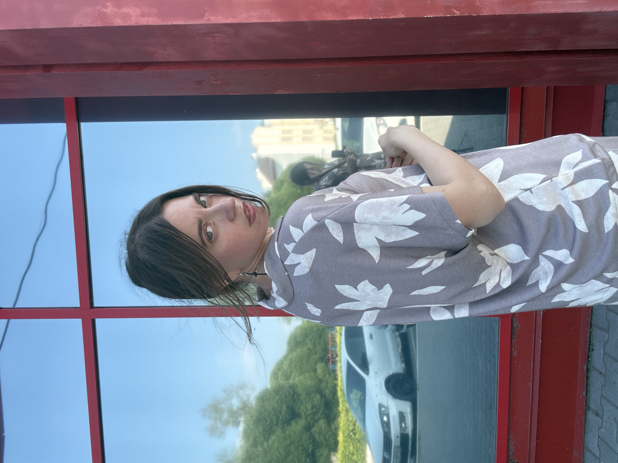 A medium shot shows a young woman with long, dark hair, wearing a gray shirt with a white floral pattern and black earrings. She is standing outside a building with red trim around a large window that reflects the sky, trees, and buildings. A white car is visible in the reflection of the window. The woman is looking over her shoulder at the camera with a serious expression. Her right hand is touching her left arm, and her left hand is raised slightly. The background is out of focus, showing a city environment.
